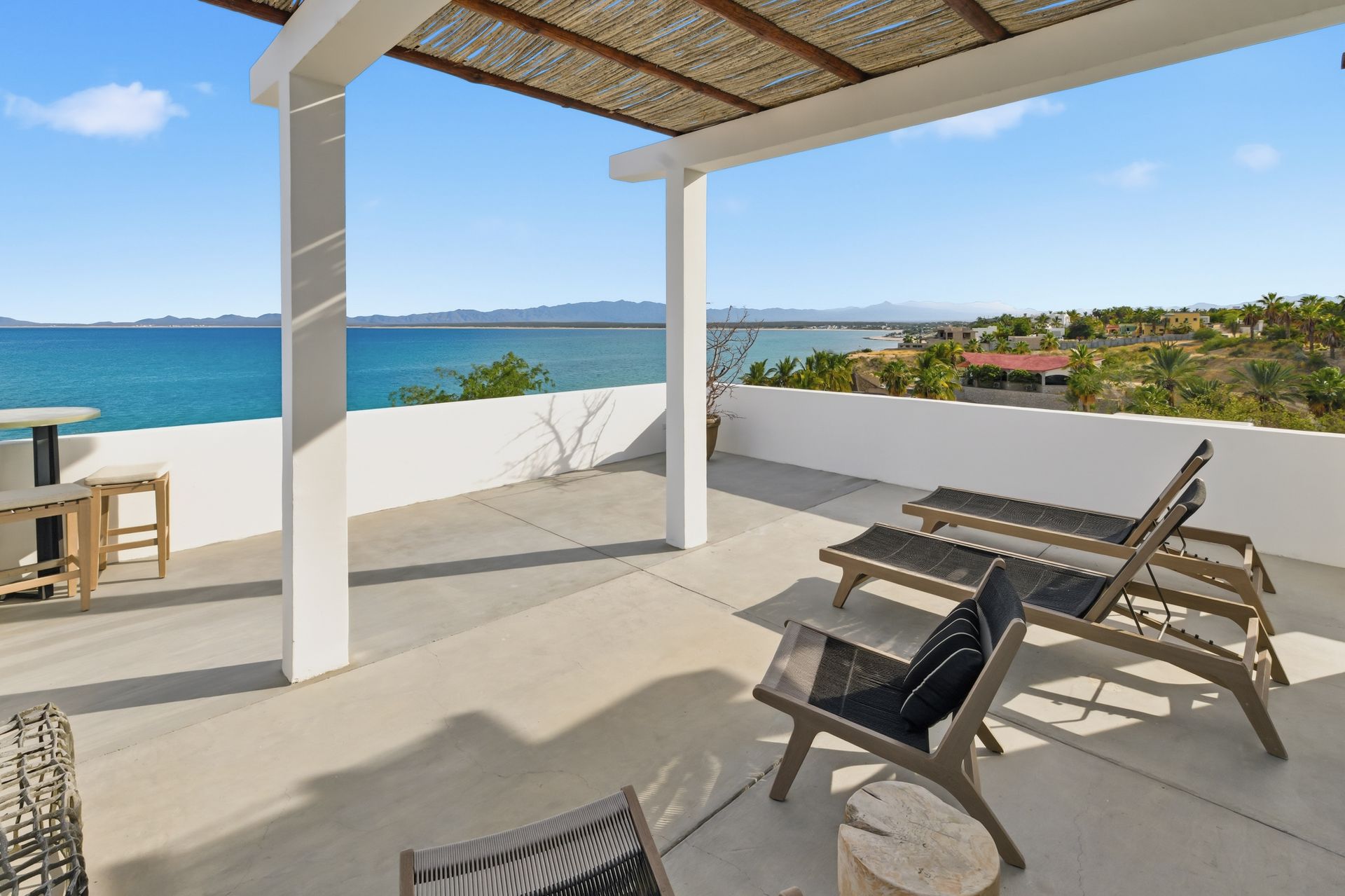 Rooftop patio overlooking ocean with lounge chairs, white columns, and straw roof.