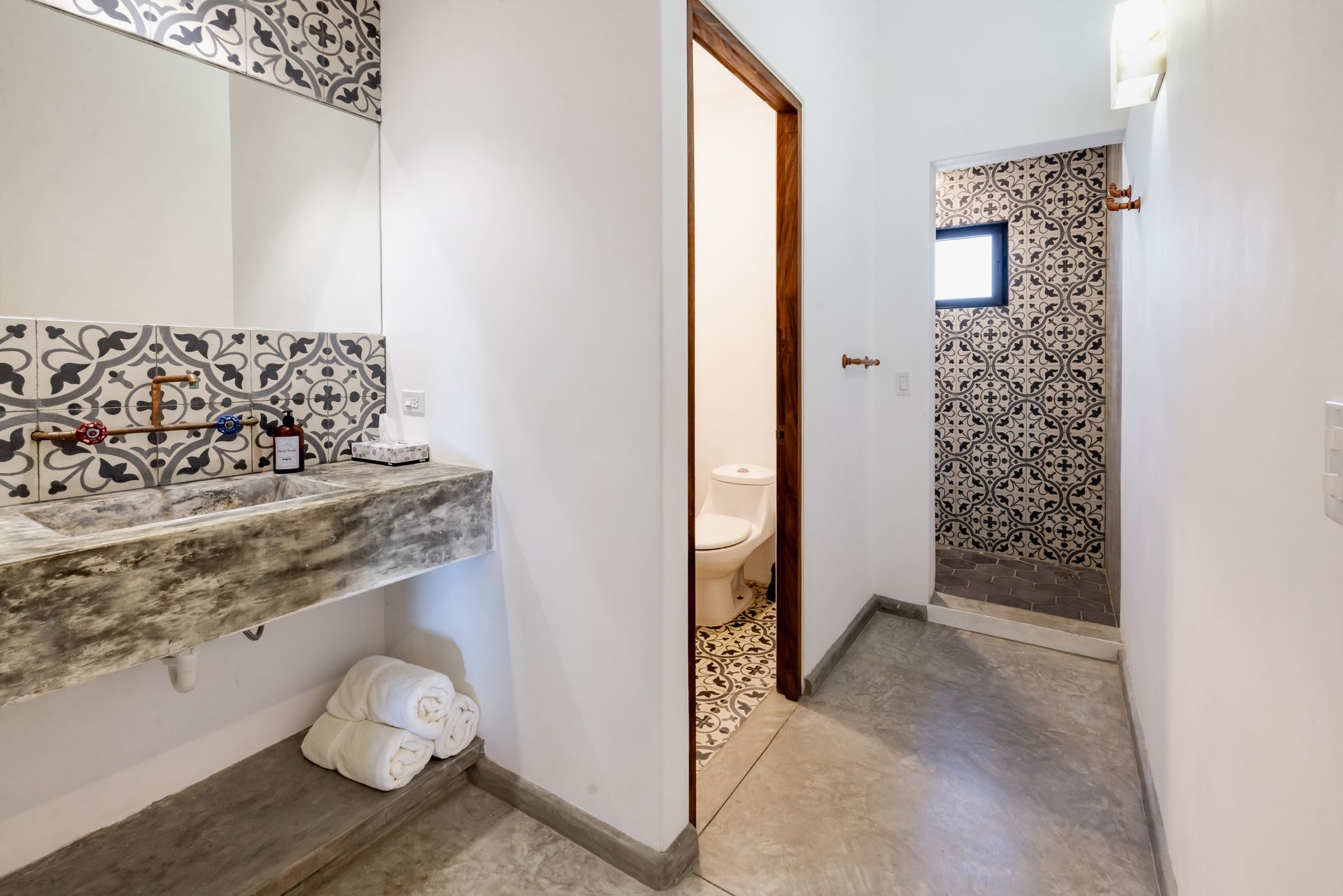Bathroom with patterned tile, concrete sink and floor, shower with a small window, and open doorway.
