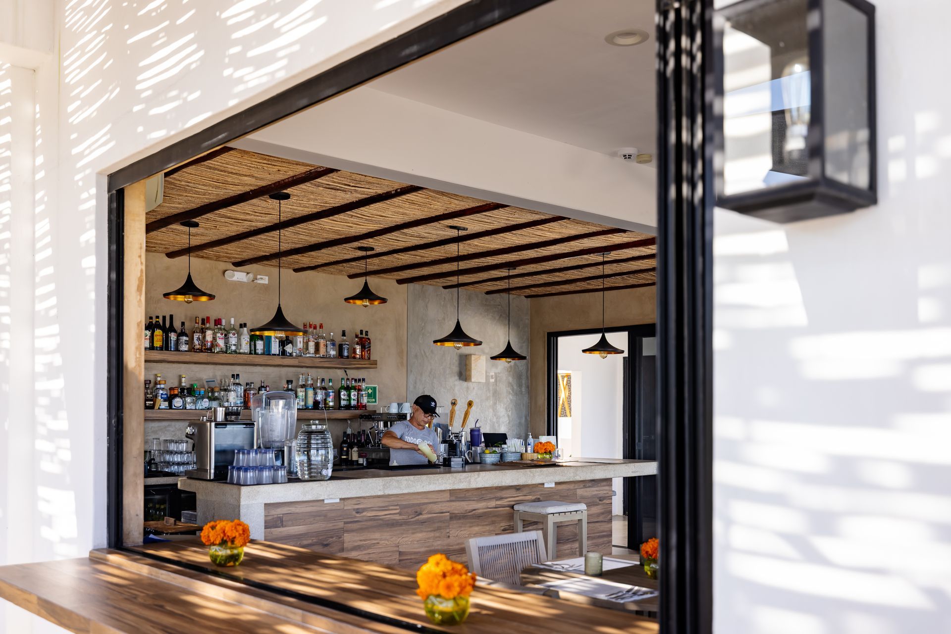 Bar with bartender preparing drinks. Open window view, white exterior, neutral tones.