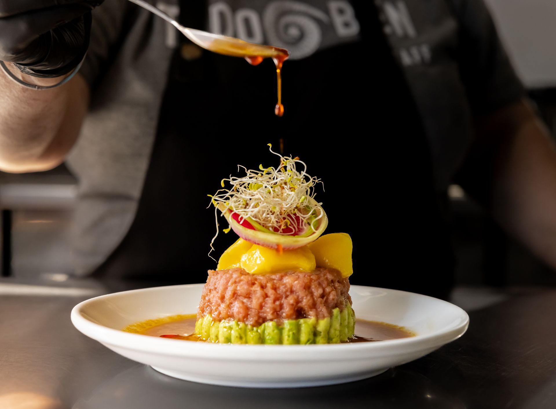 Chef plating a stacked dish of avocado, salmon, and mango with sauce.