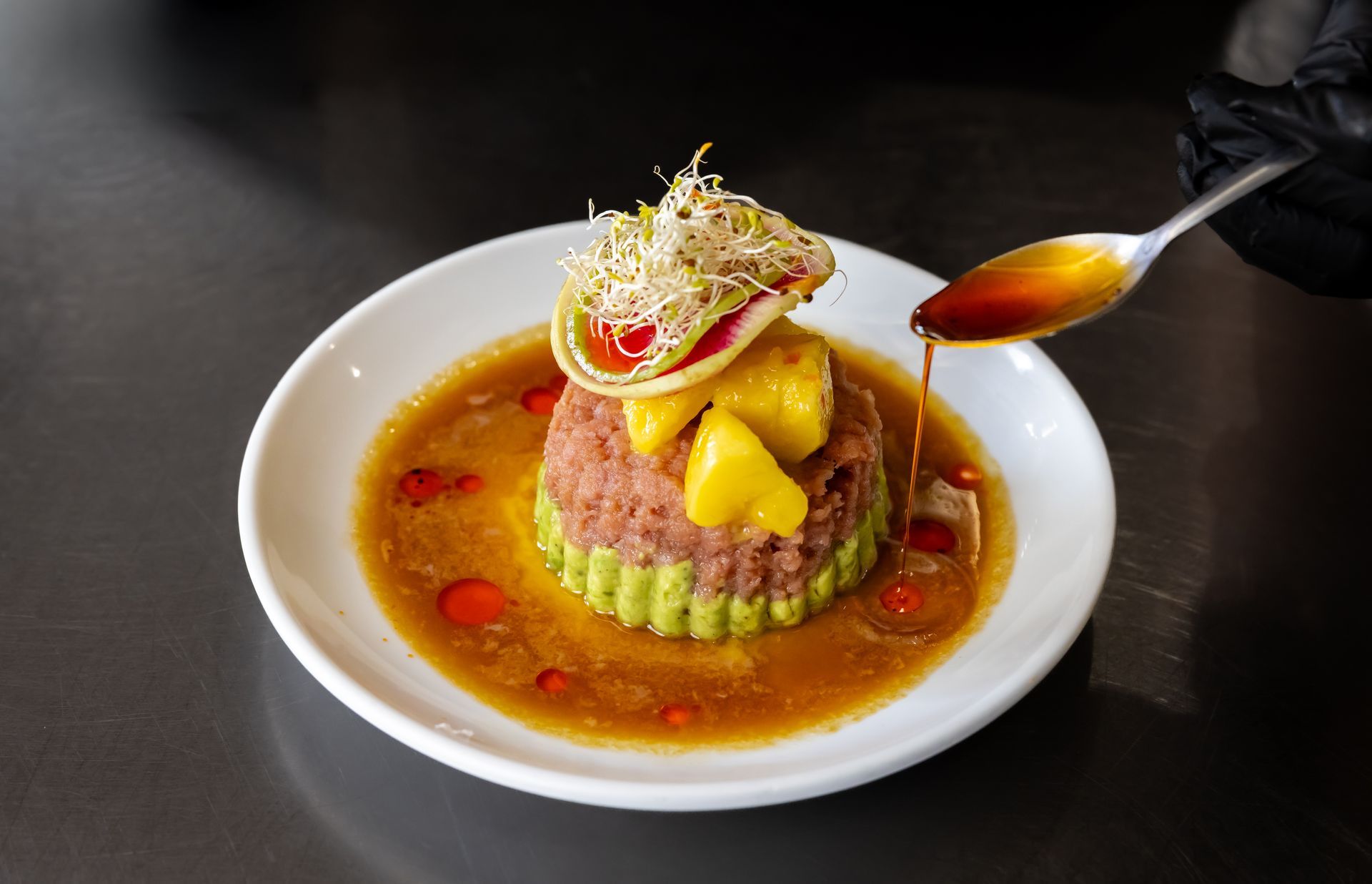 Gourmet dish: Stacked food with sauce being drizzled. White plate on a dark table.