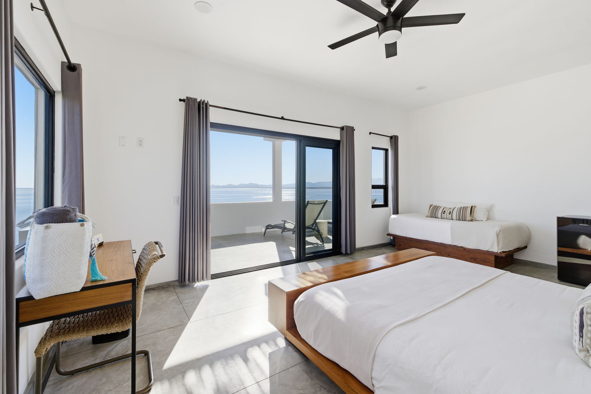 Bedroom with large bed, balcony, desk, and daybed; windows offer ocean view.
