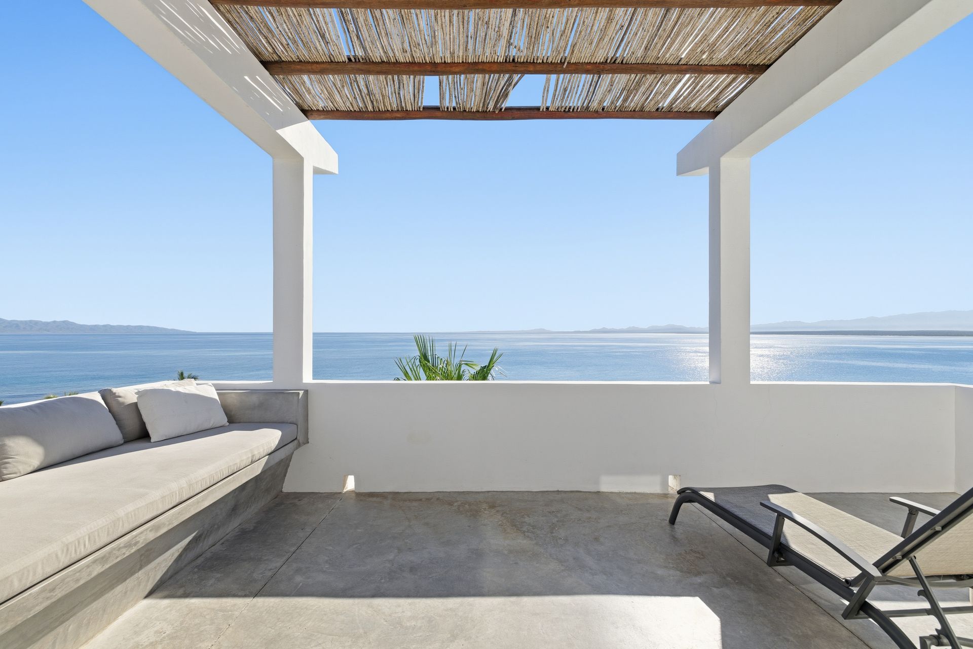 A white, open-air balcony overlooks the sea. It features a built-in couch, a lounge chair, and a woven roof.