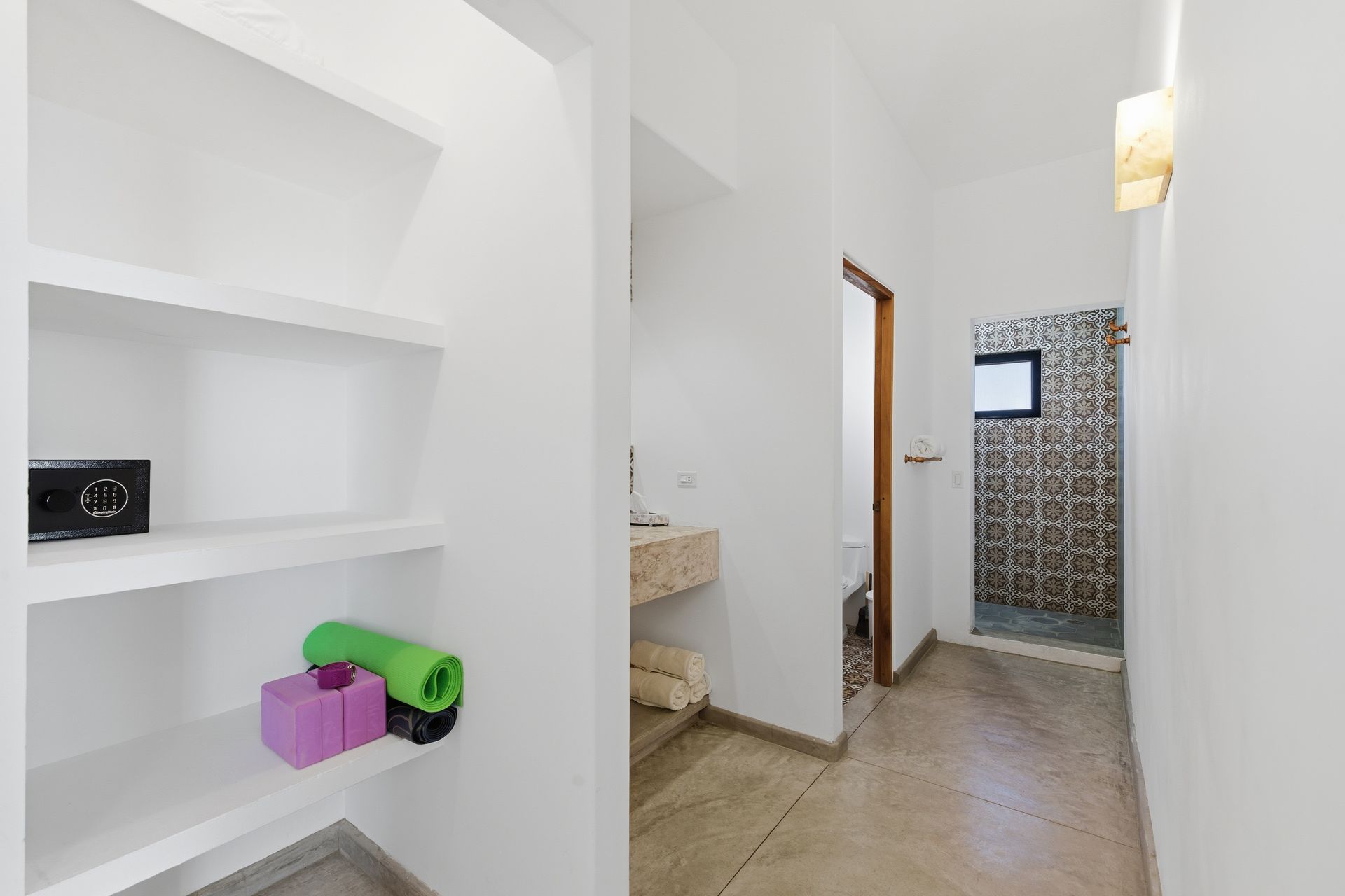White hallway with built-in shelves holding a safe, yoga mat and blocks, leading to a bathroom.