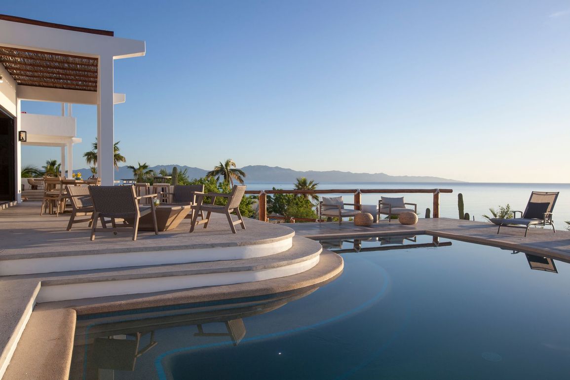 Luxury home with infinity pool overlooking ocean and mountains; chairs on patio.