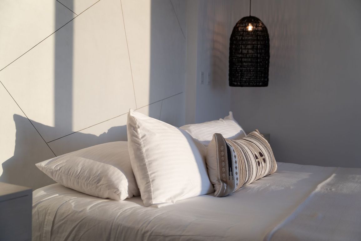 Bedroom with white bedding, pillows, and a decorative woven light fixture; sunlight on the wall.