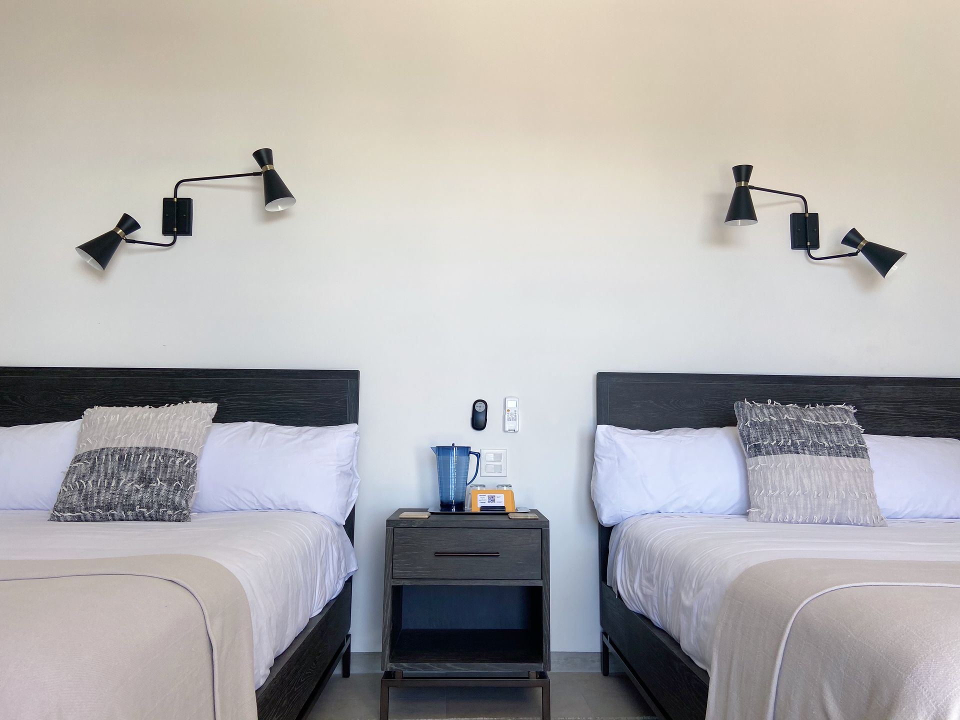 Two beds with white linens, flanked by black wall lamps. Nightstand with small pitcher and outlet.