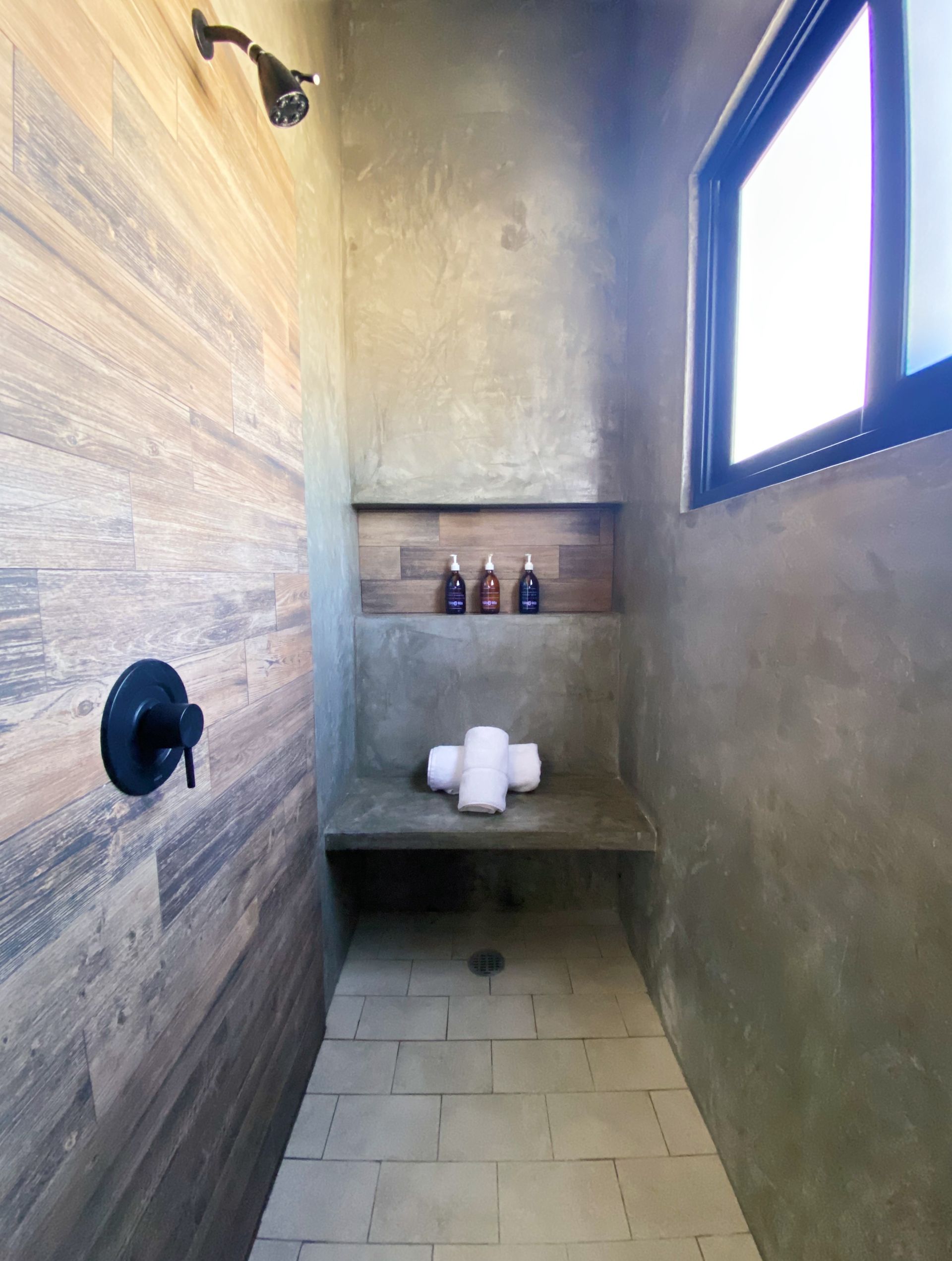 Shower with wood-look tile on one wall, a concrete-look wall with a shelf and small window, and dark fixtures.
