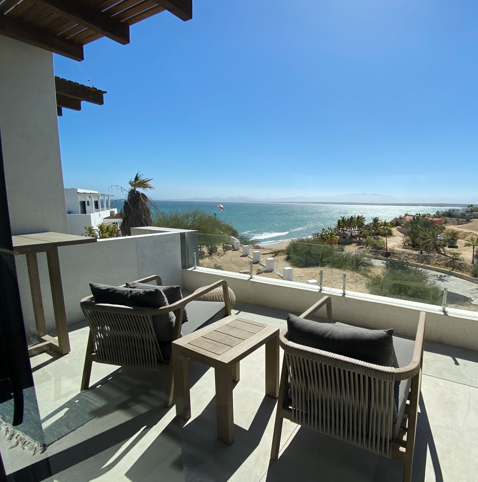Balcony with ocean view. Two gray cushioned chairs, small table, clear railing, sunny day.