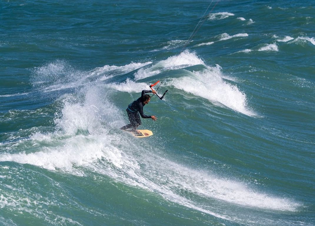 Person kitesurfing on a wave, ocean. Wearing dark wetsuit, riding on a board, with a kite above.