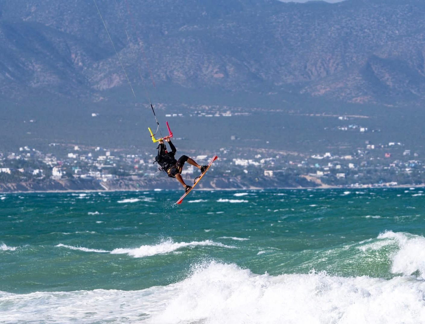 Kitesurfer airborne over ocean waves, kite overhead. Mountains and town in background.