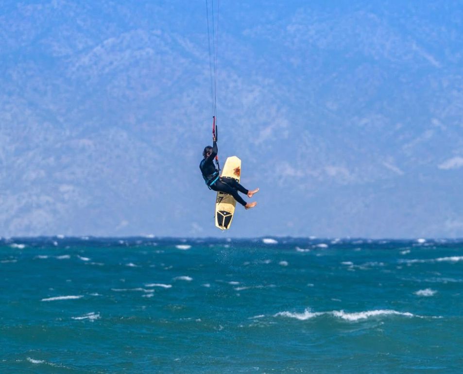 Kitesurfer in mid-air, holding on, over choppy blue water. Mountains in background, under a blue sky.