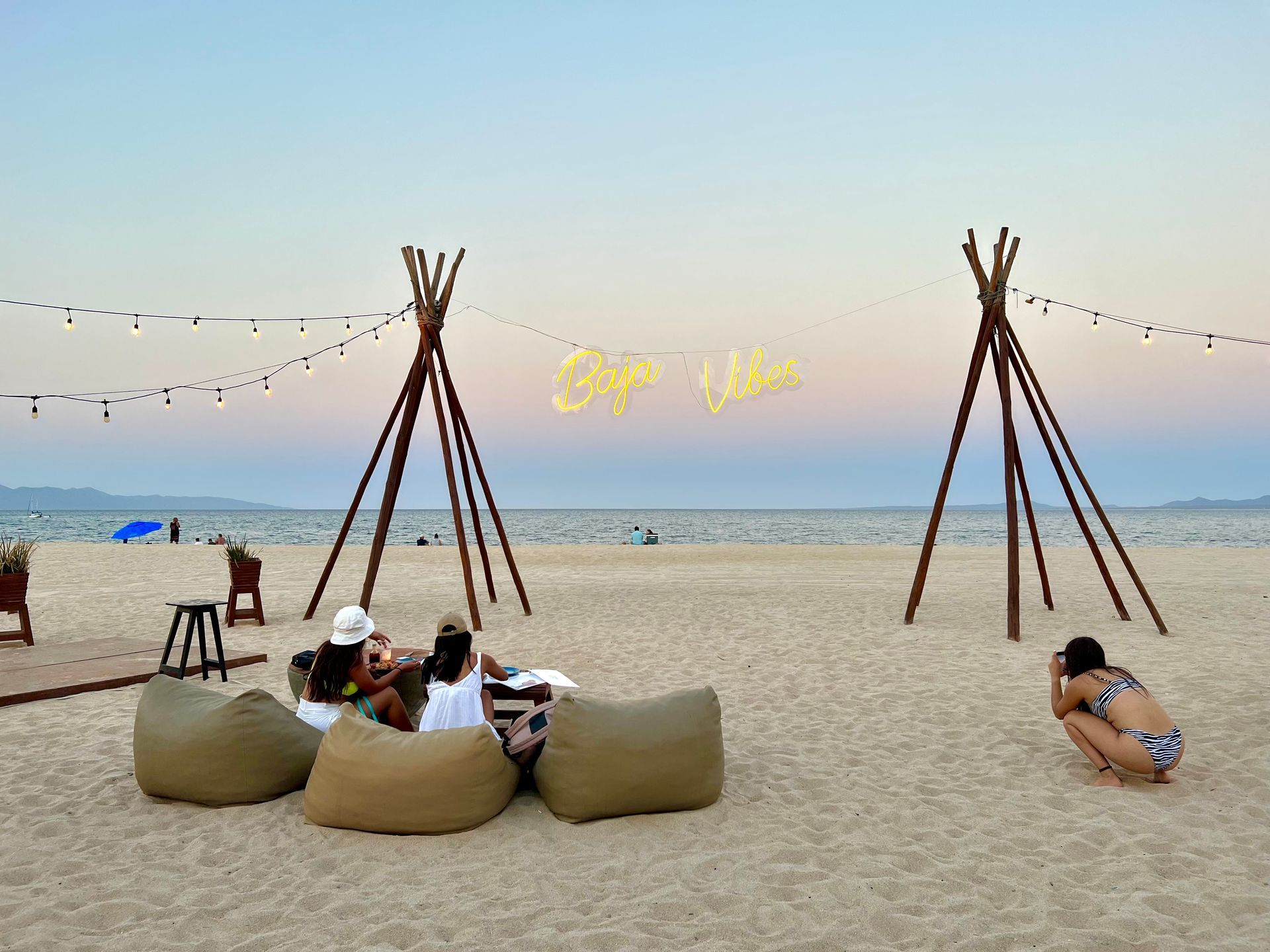 Beach scene with beanbag chairs, teepee-like structures, string lights, and people relaxing by the water.