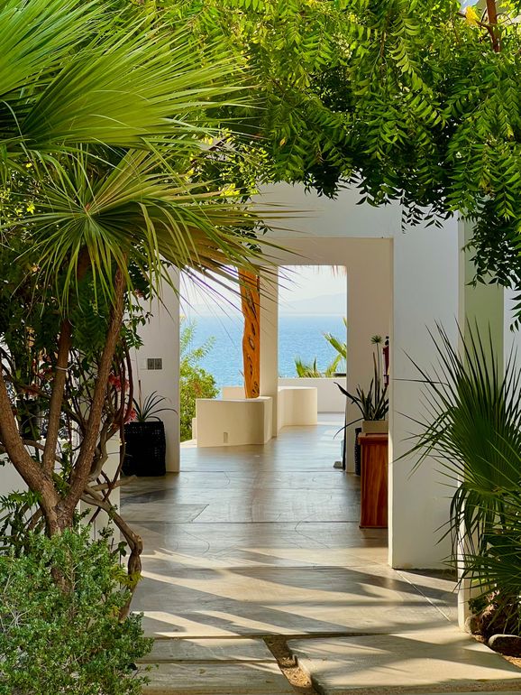 Open-air white hallway with ocean view, framed by green plants and trees.