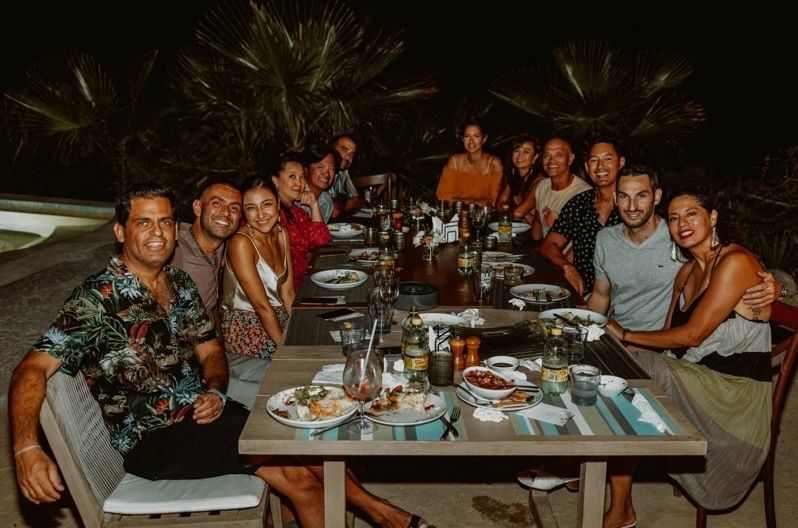 A group of people sitting around a long outdoor table at night, eating and smiling.