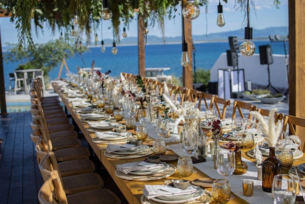 Long wooden table set for an outdoor event, overlooking the ocean; adorned with flowers and glassware.