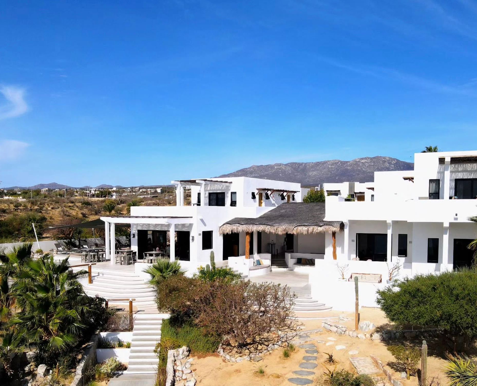 White villas with dark windows and palm-thatched roof, set on a desert landscape under a blue sky.