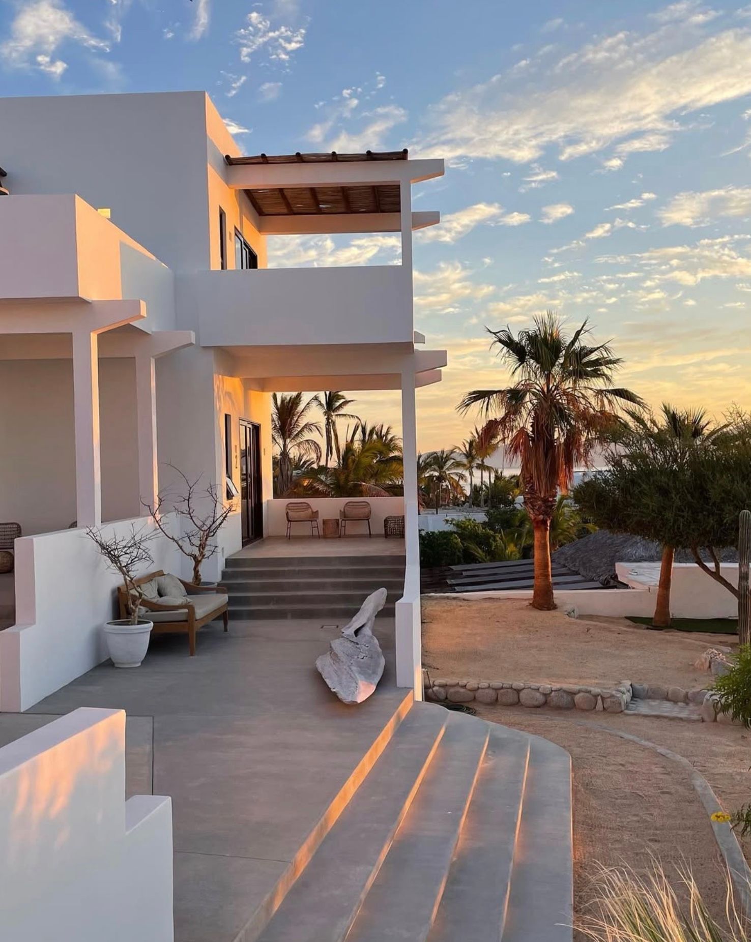 White two-story building with a terrace and stairs, palm trees, and a sunset in the background.