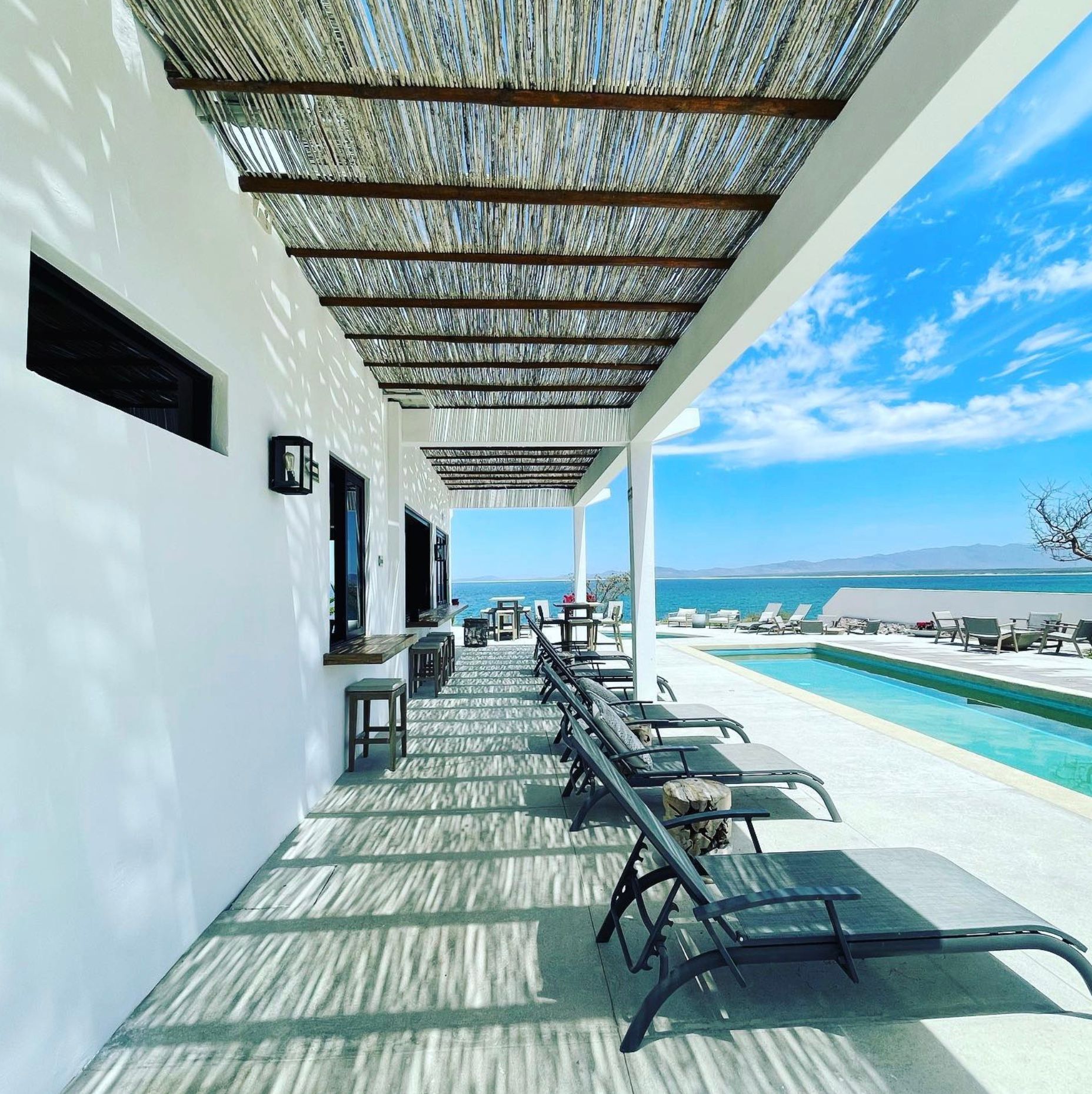 A row of lounge chairs beside a pool and ocean, under a shaded patio.