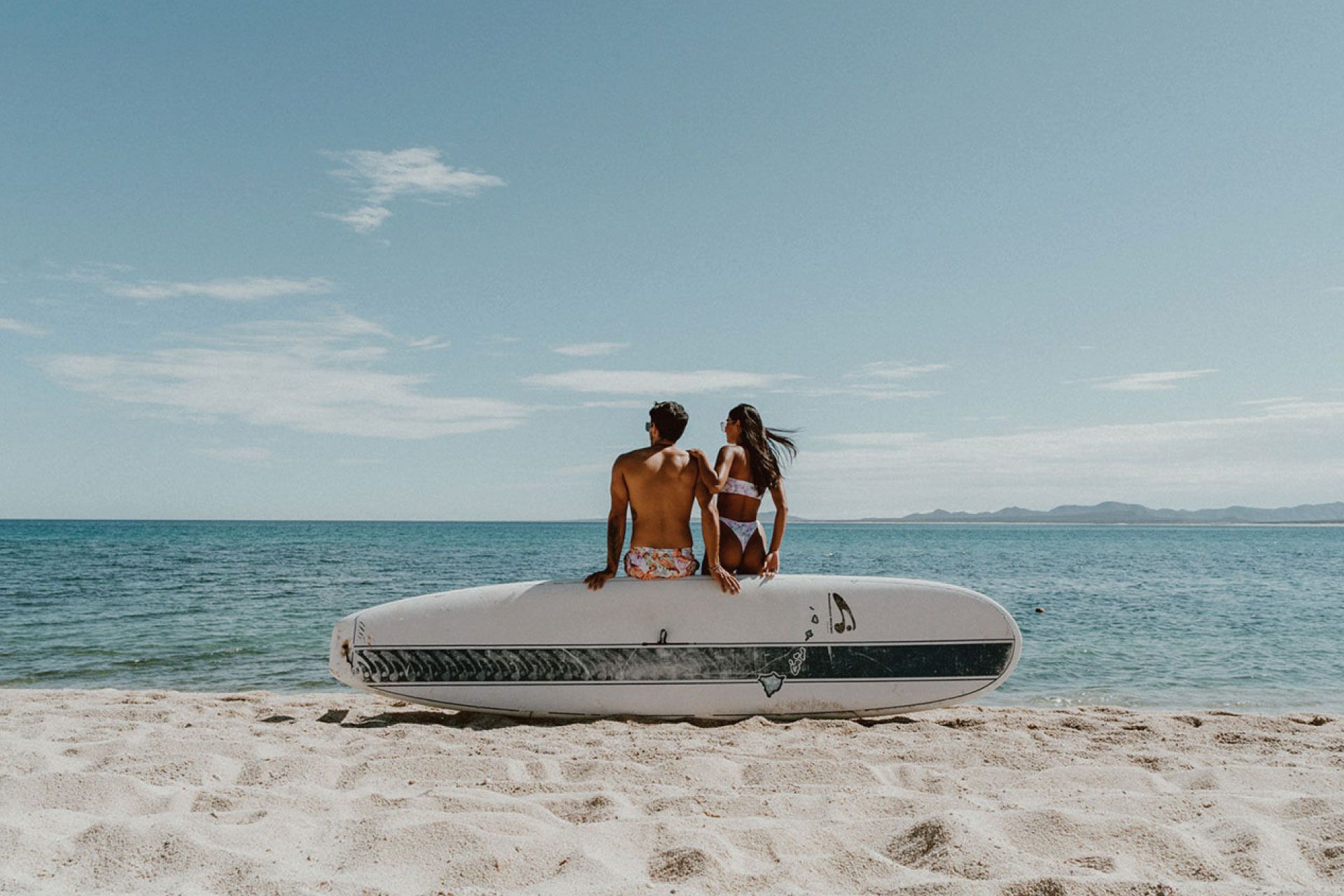 Couple sitting on a surfboard on a sandy beach, gazing at the ocean under a blue sky.