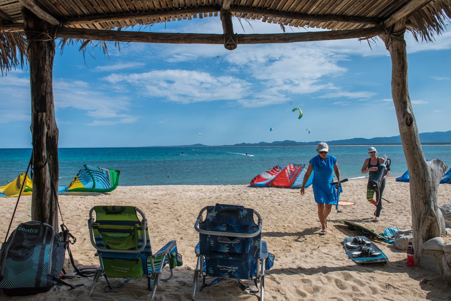 Beach scene with kitesurfers, blue sky, and thatched shelter.