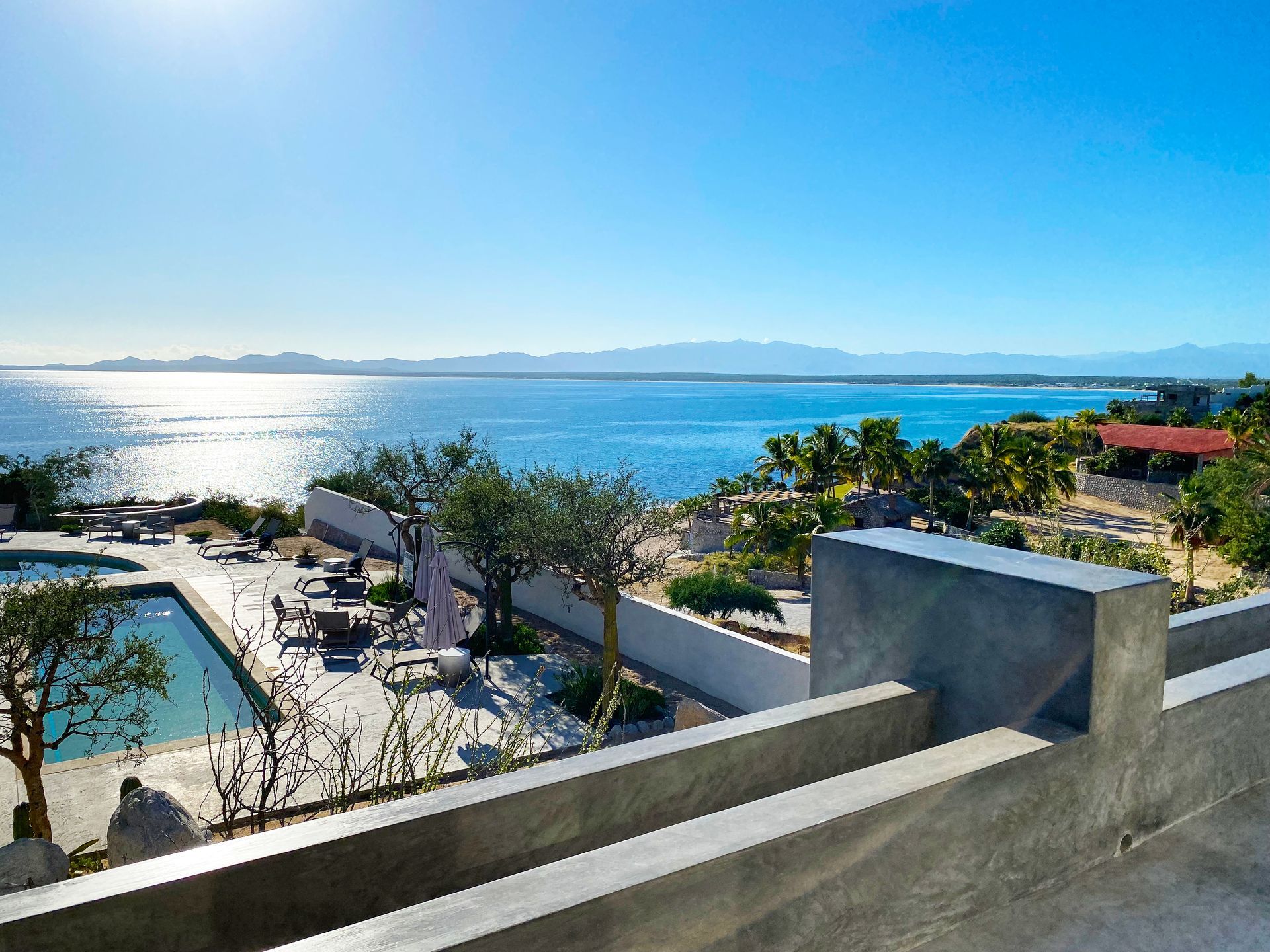 View of ocean and a pool from a concrete patio; clear blue sky, sunny day.