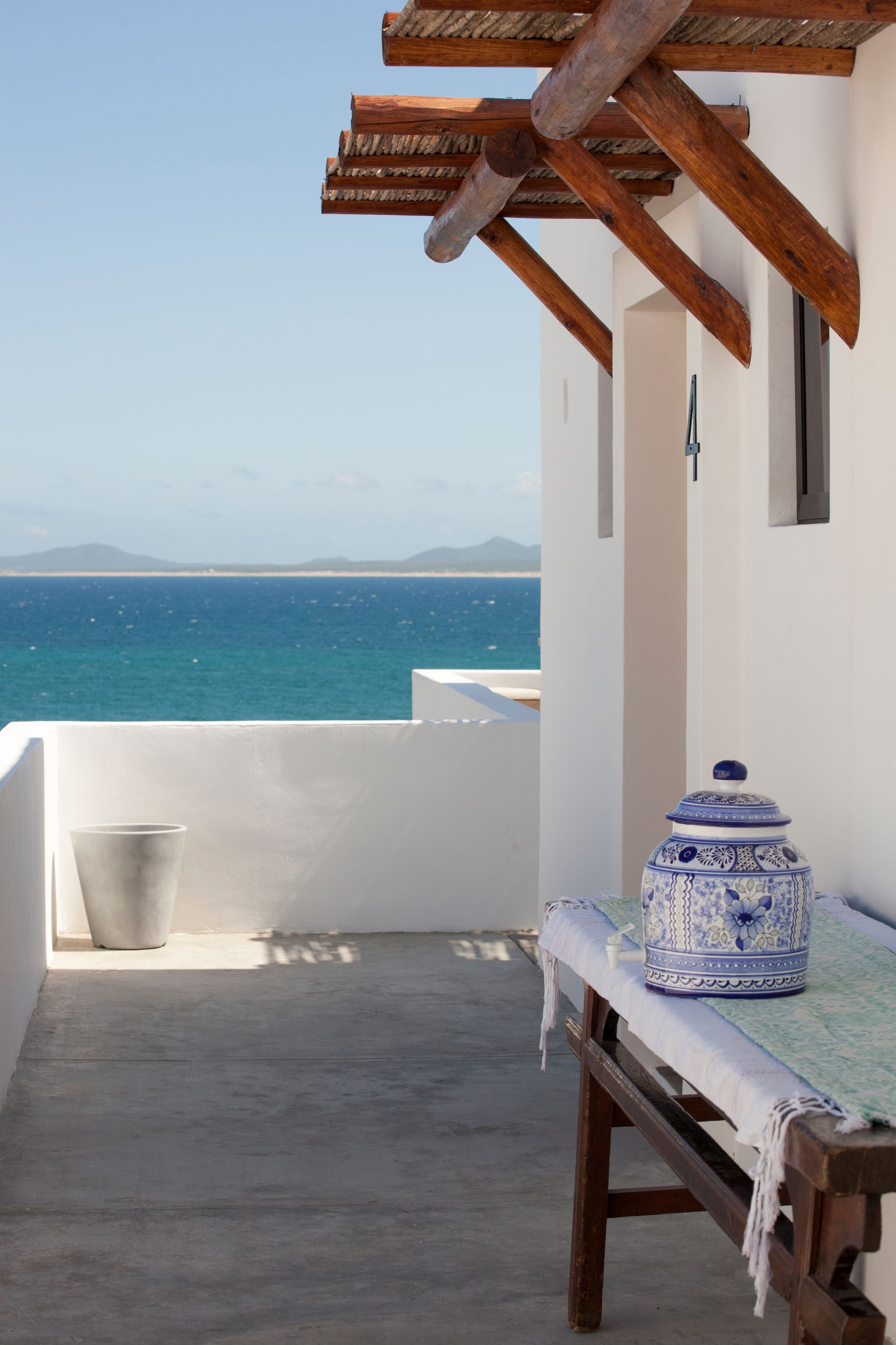 Coastal balcony with white walls, ocean view, wooden beam awning, and blue and white jar.