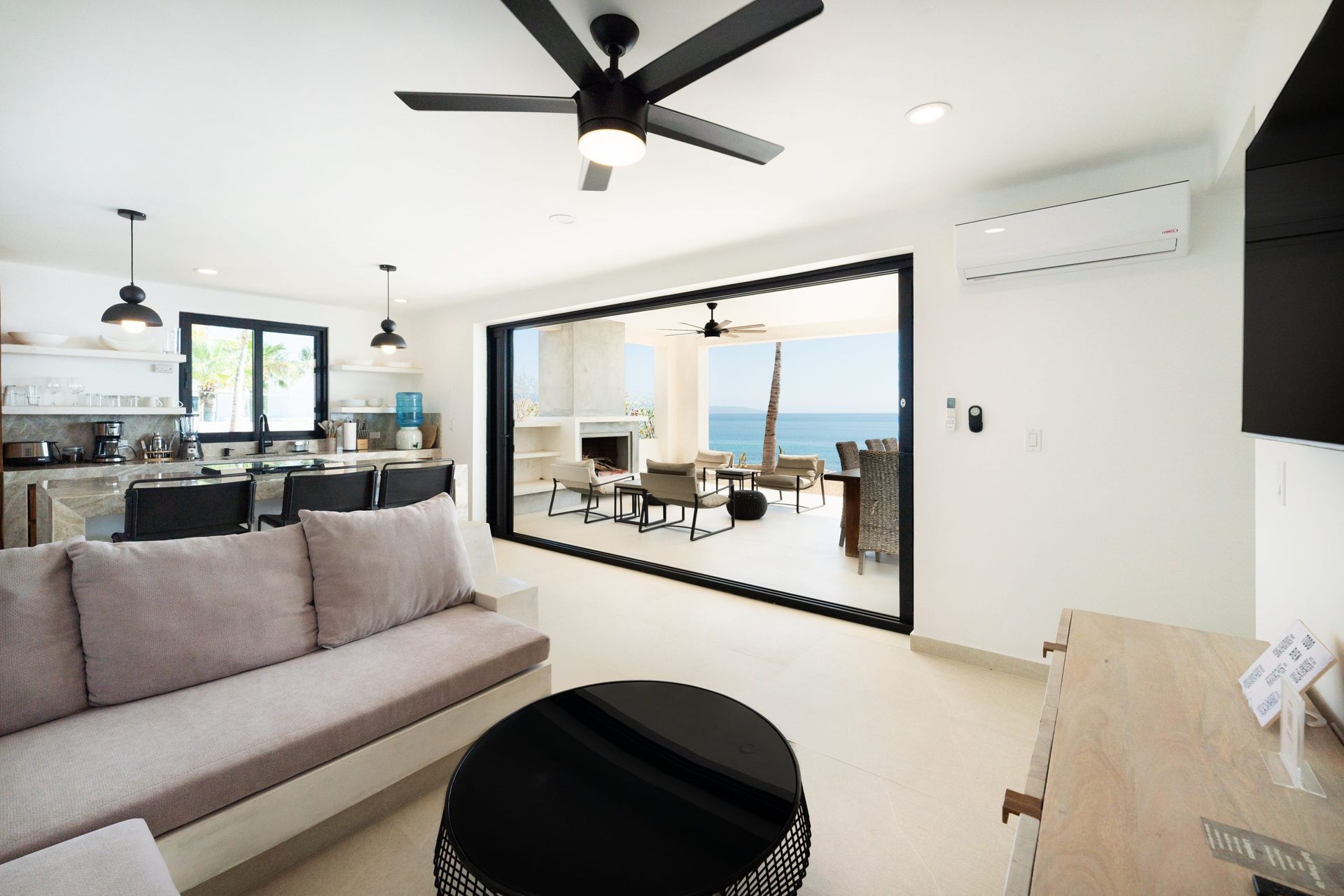Living room with open doors to ocean view; gray sofa, black coffee table, open kitchen.