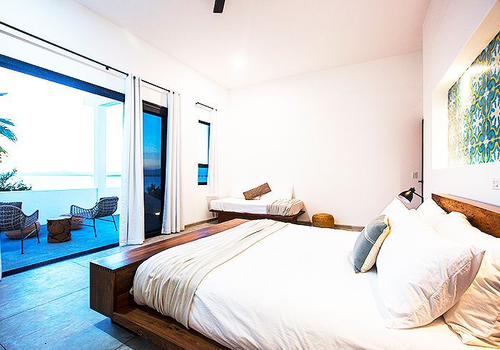 Bedroom with a bed, open doors to a patio overlooking the ocean. White bedding, wooden furniture, and neutral tones.