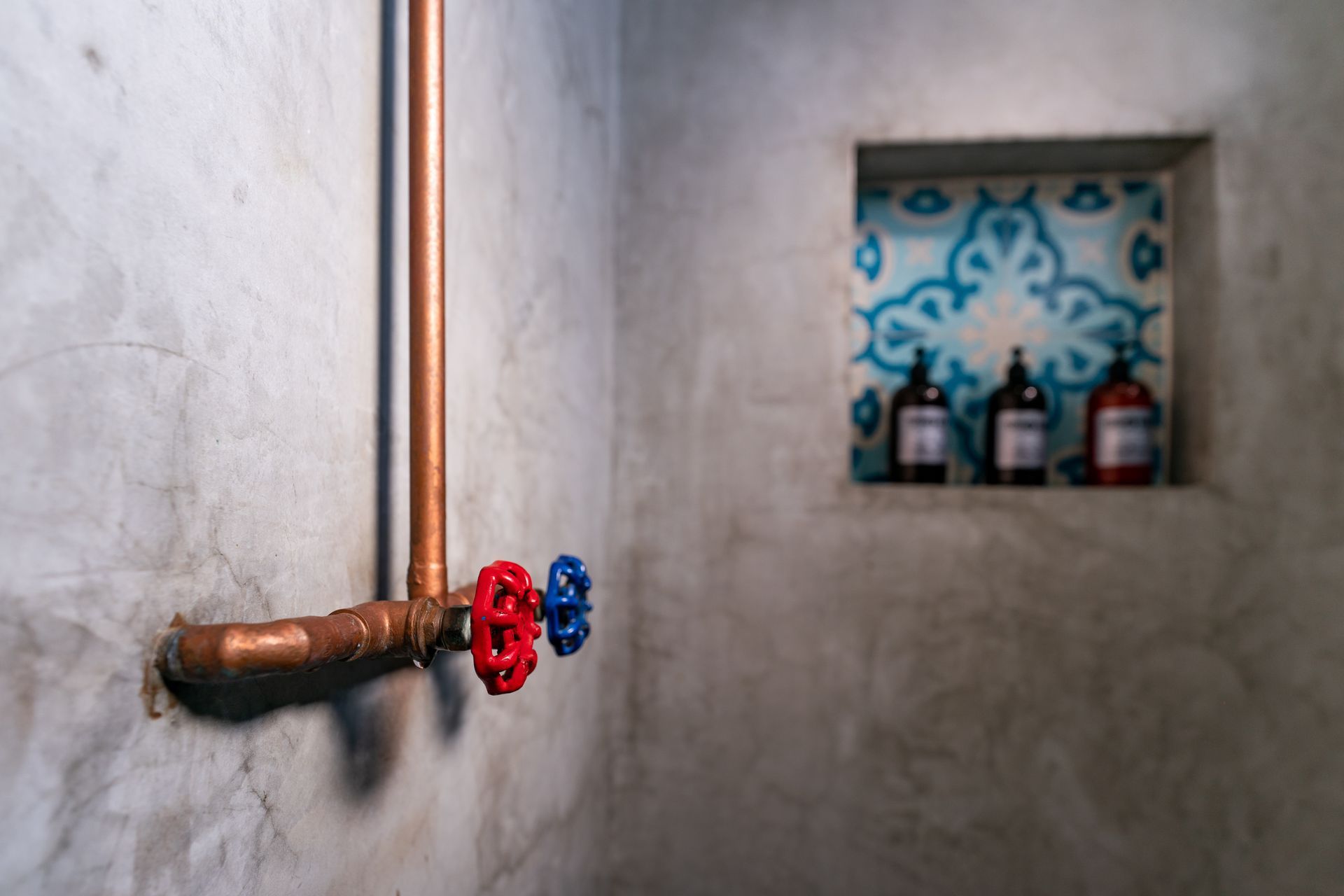 Copper shower pipes and valves on a gray concrete wall, with a tiled alcove holding bottles.