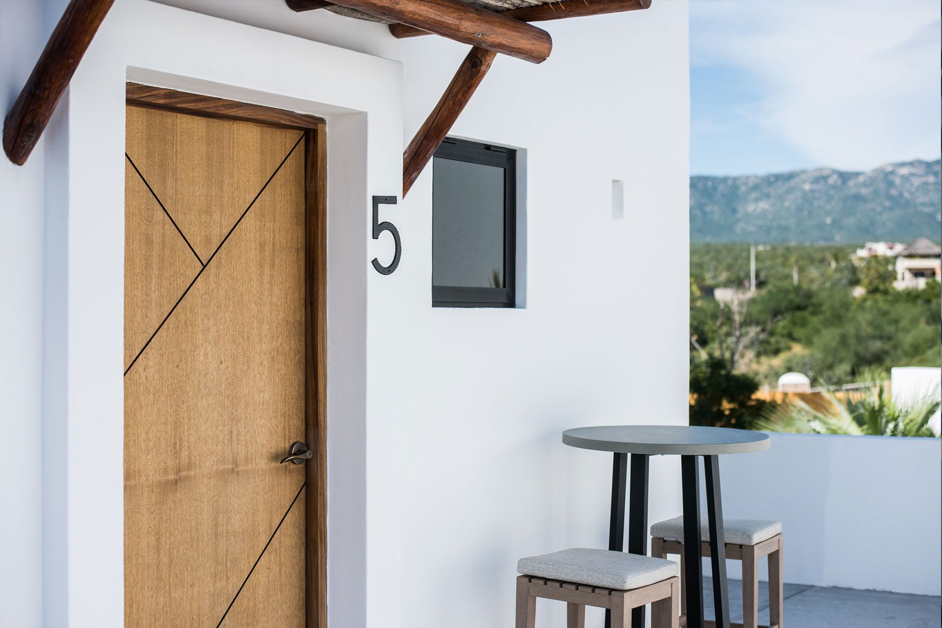 Exterior of a room with a wooden door, number 5, and a small table with stools on a white patio.