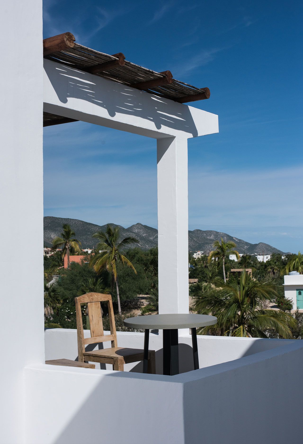 Balcony with white pillars, table, and chair. Lush green trees and mountains in the distance against a blue sky.