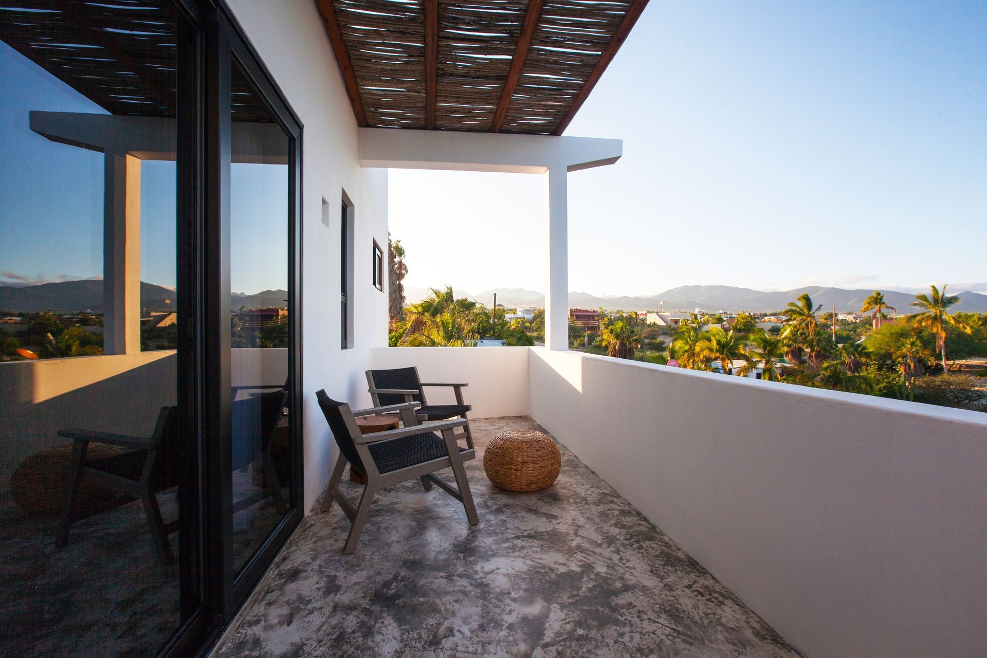 Balcony with chairs and woven ball, overlooking a scenic view of palm trees and mountains under a clear sky.