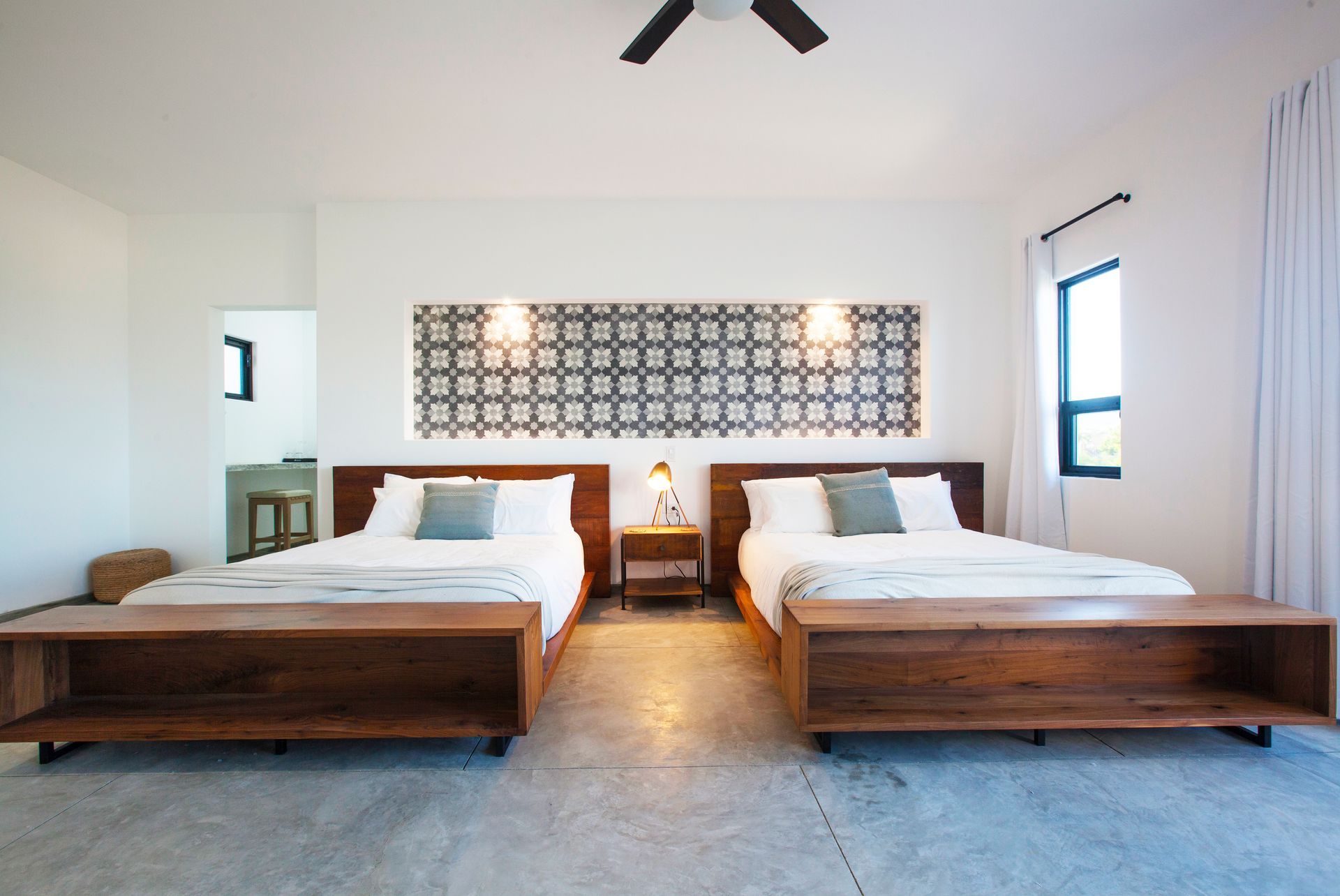 Two beds in a bright room with patterned wall decor, a small nightstand, and a window.