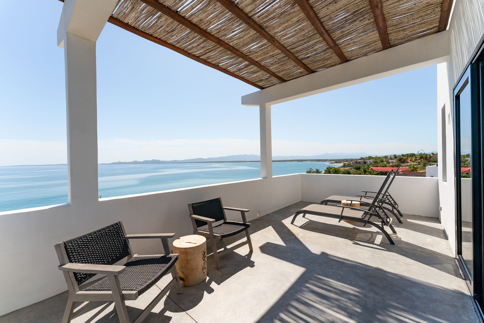 Balcony overlooking ocean, featuring chairs, sunbeds, and a thatched roof.