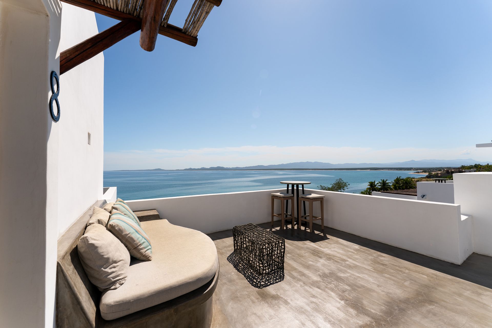 Rooftop patio with ocean view, round sofa with pillows, small table and stools, bright sunny day.