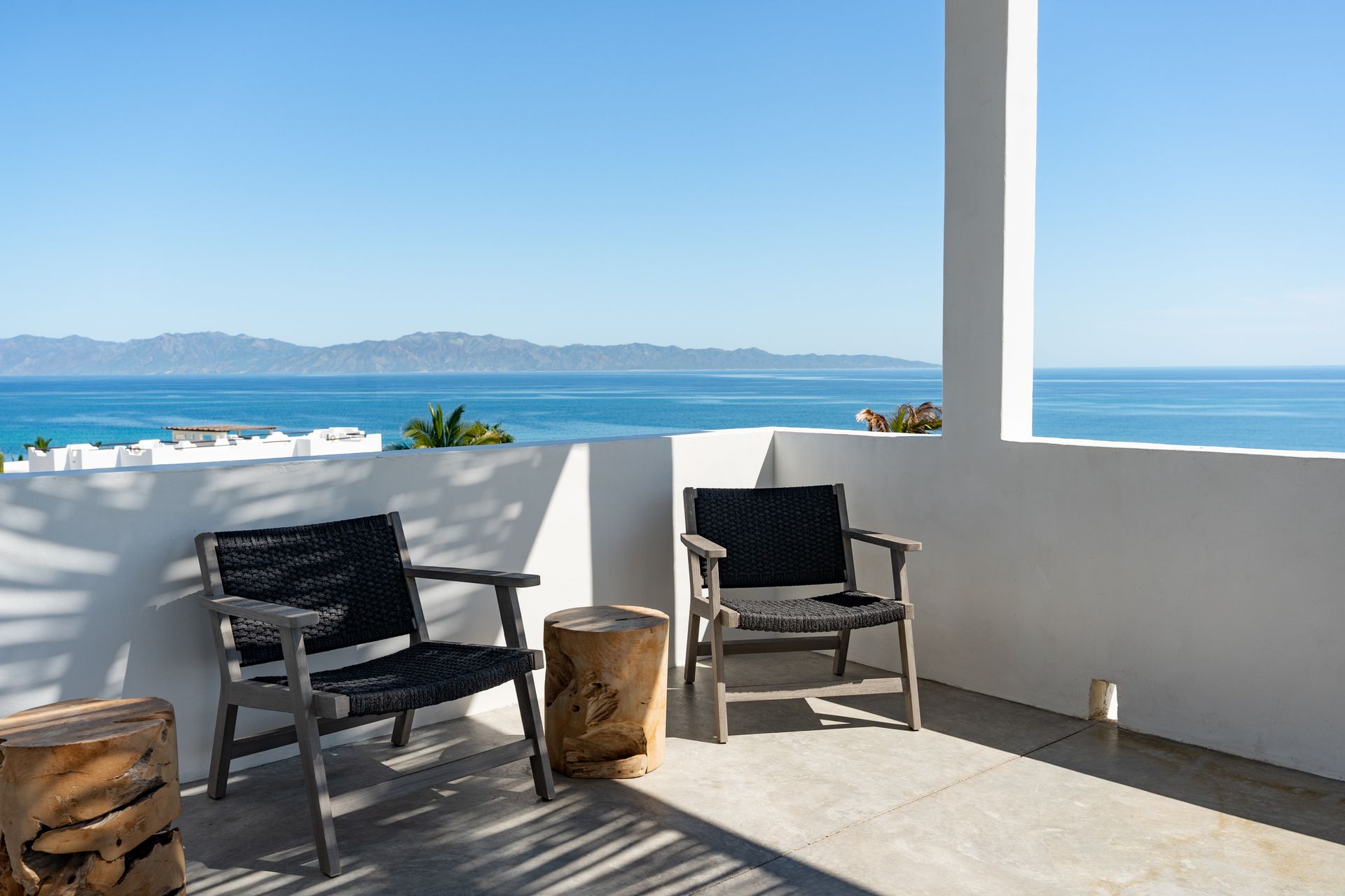 Two chairs on a white balcony with ocean view, blue sky and mountains in the distance.