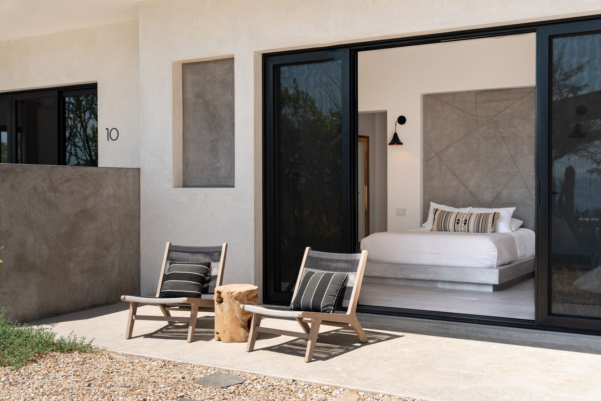 Patio with two chairs and a small table, leading to a bedroom with bed and sliding glass doors.