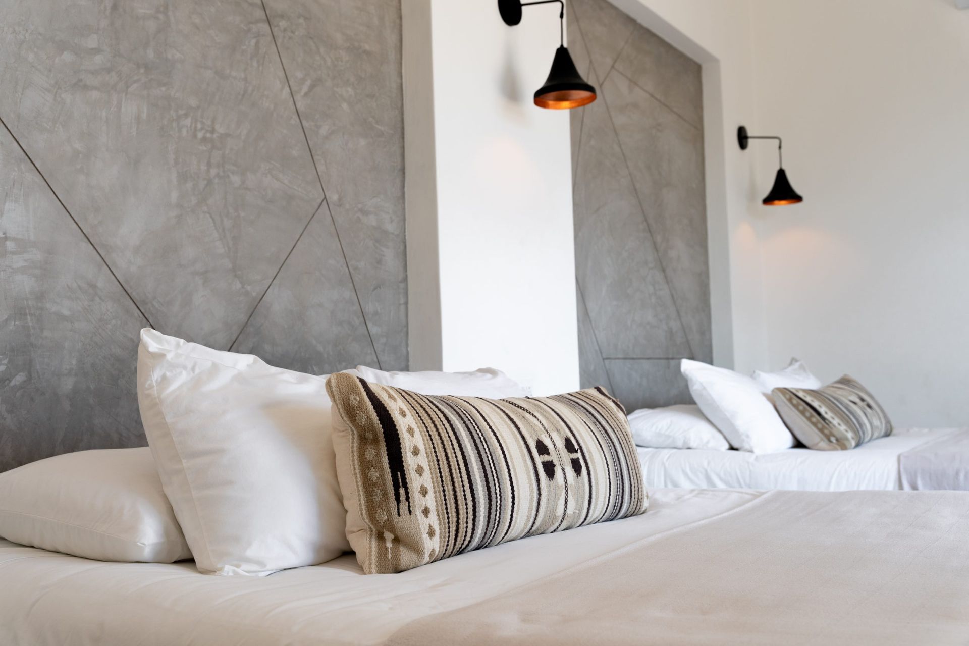 Two beds with white bedding, patterned pillows, against concrete and white walls, sconce lighting.