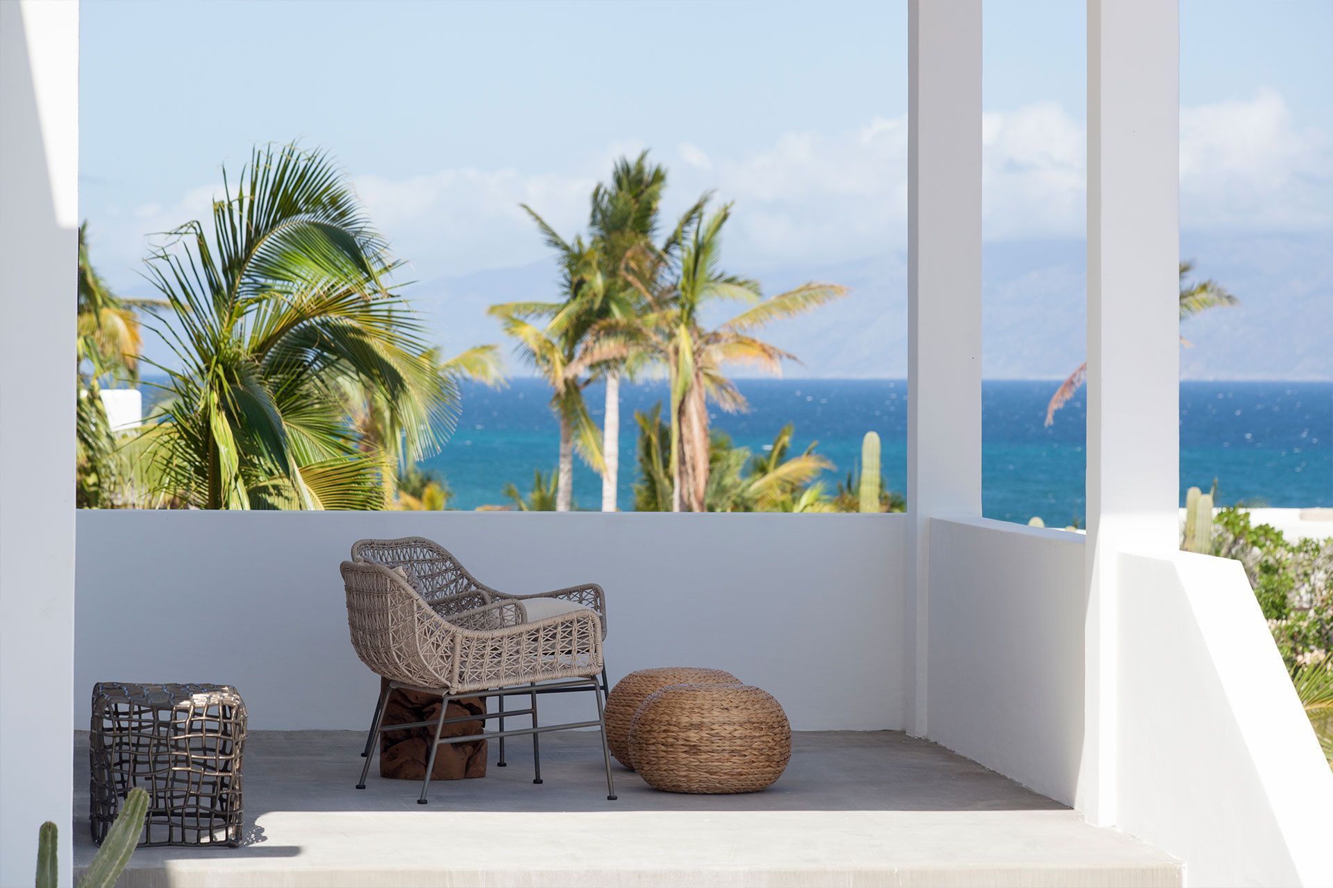 Patio with woven chair, footstool, and view of palm trees and ocean.