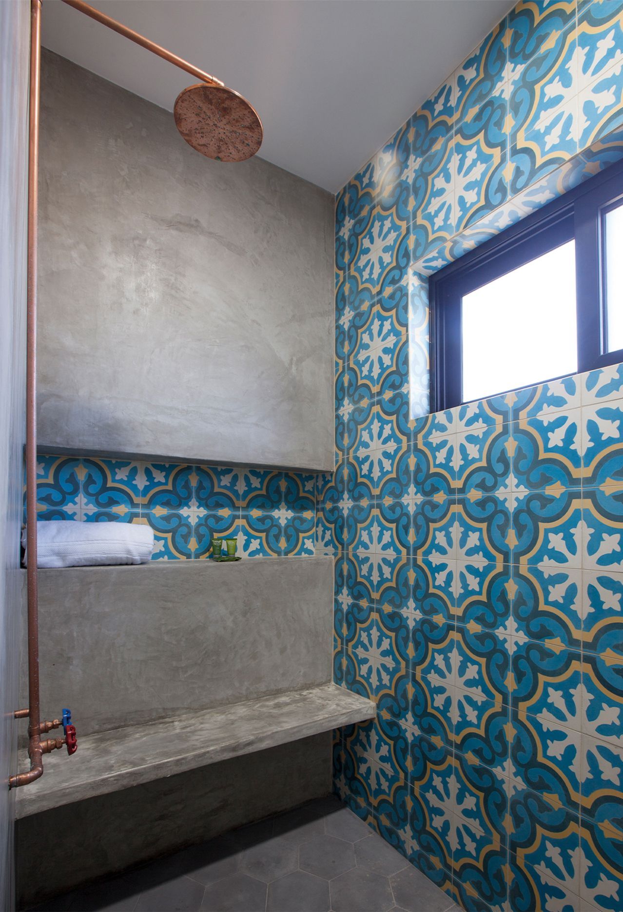 Shower with blue and white patterned tile and copper fixtures. Concrete bench and gray wall.
