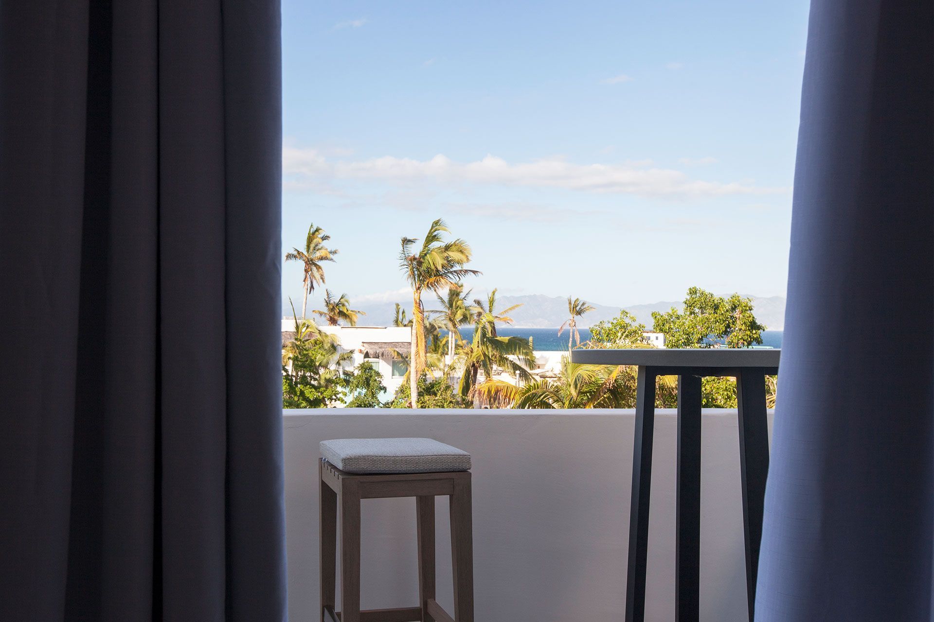 Balcony view of the sea and palm trees framed by curtains, with a stool and small table.