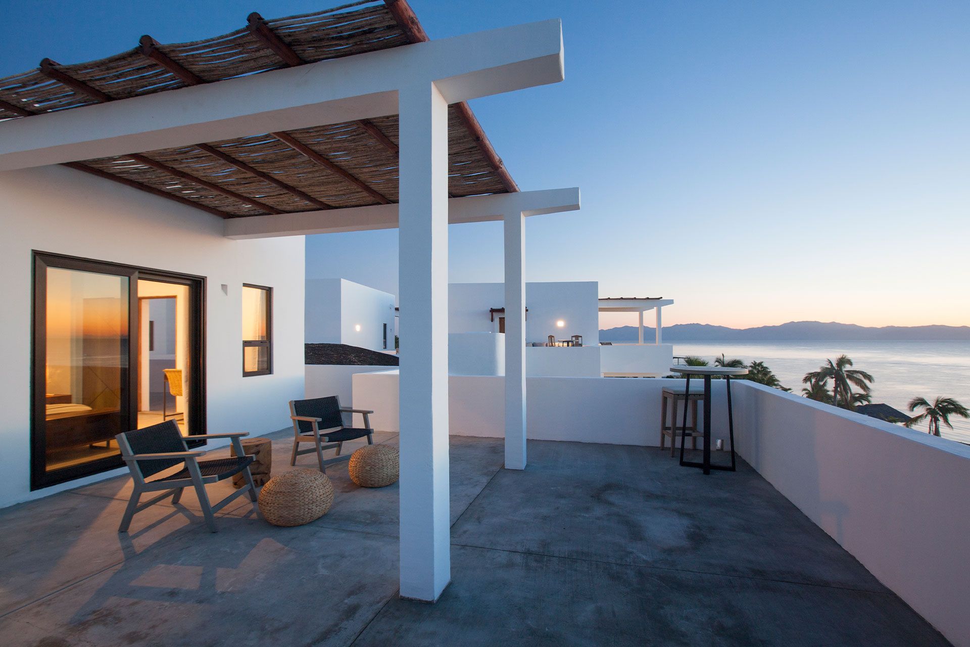 Rooftop patio overlooking ocean at dusk. White pillars support a woven shade. Two chairs, two poufs, and a high table.