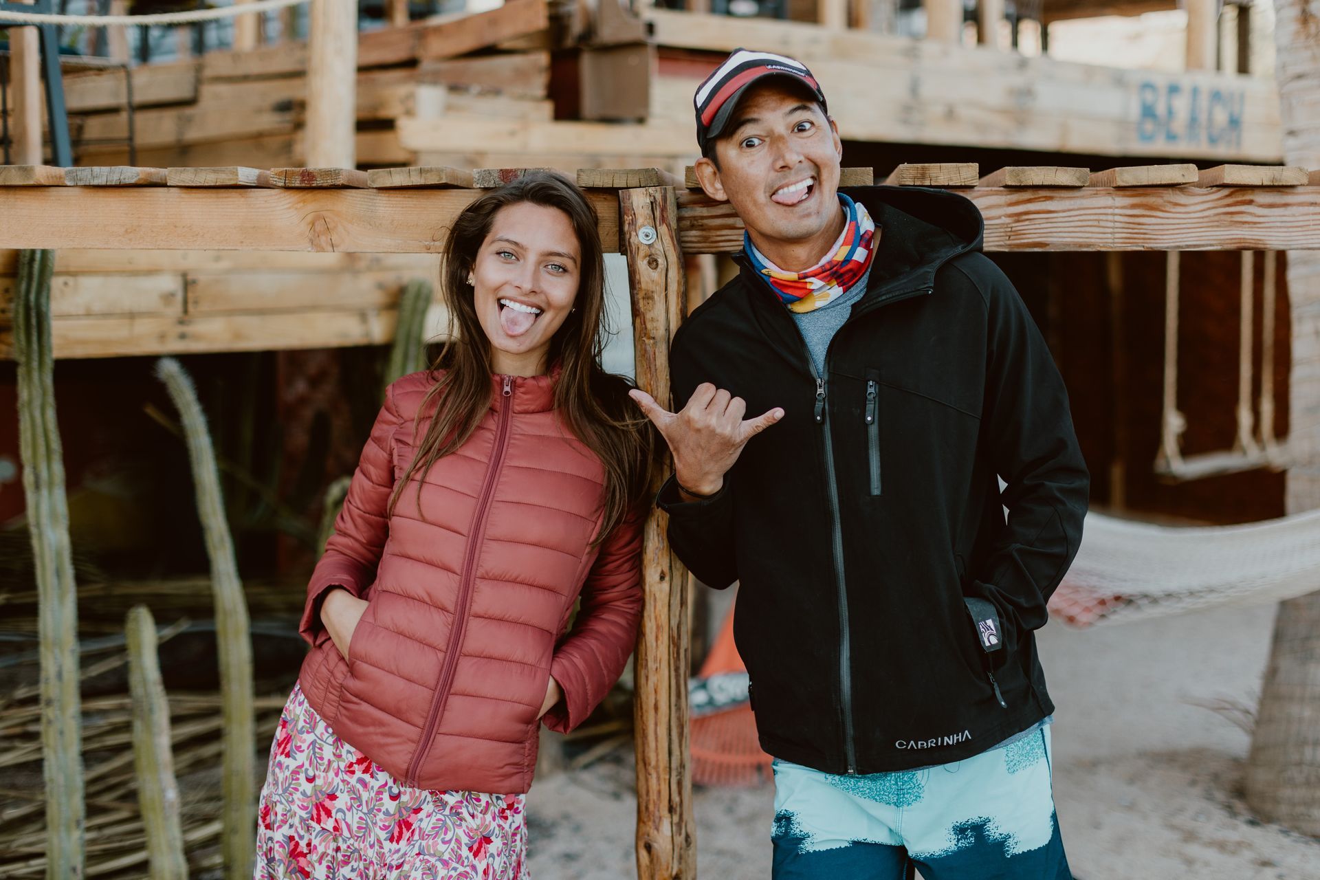 Two people making funny faces near a wooden structure on a sandy beach.
