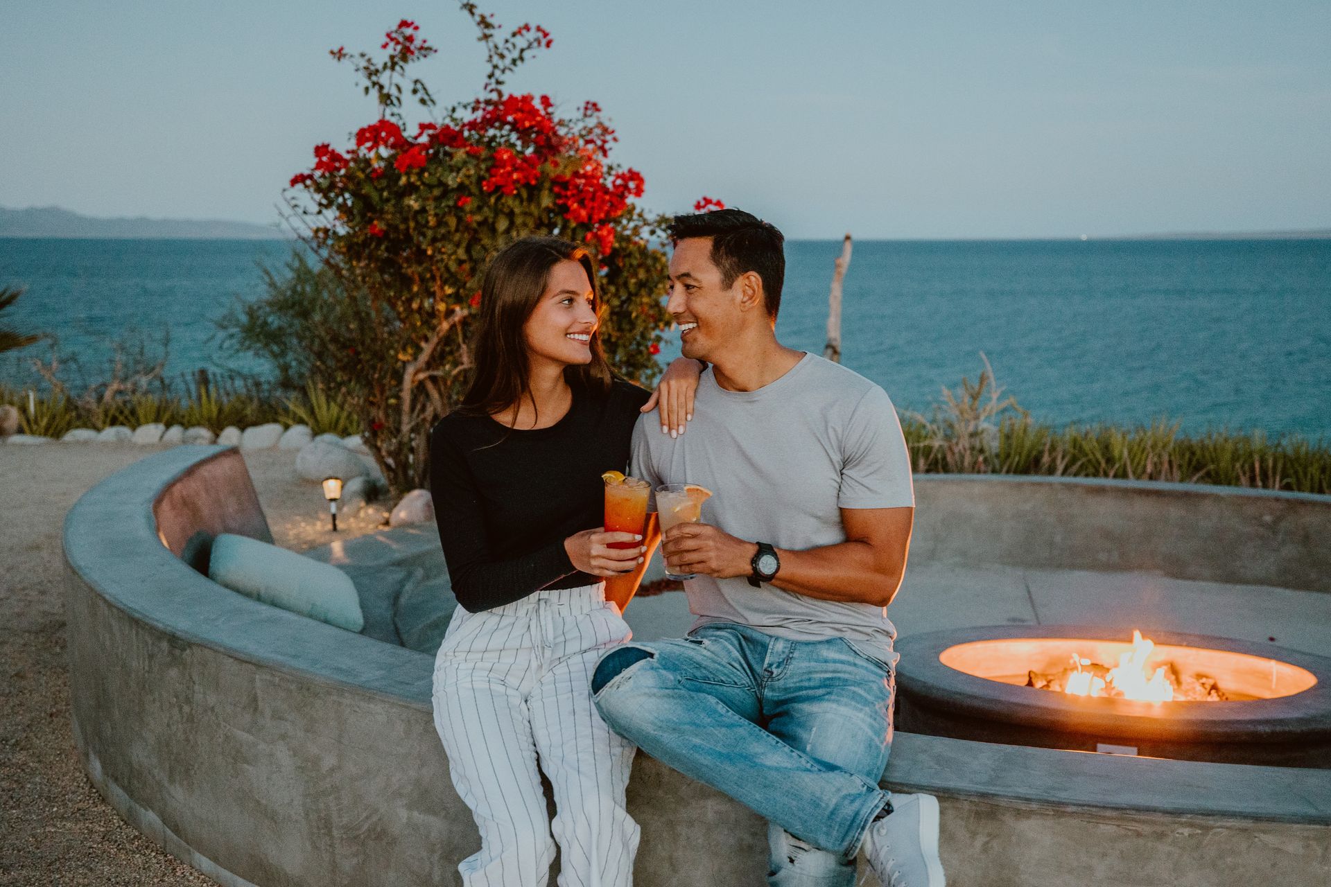 Couple seated outdoors with drinks, looking at each other. Fire pit and ocean view in background.