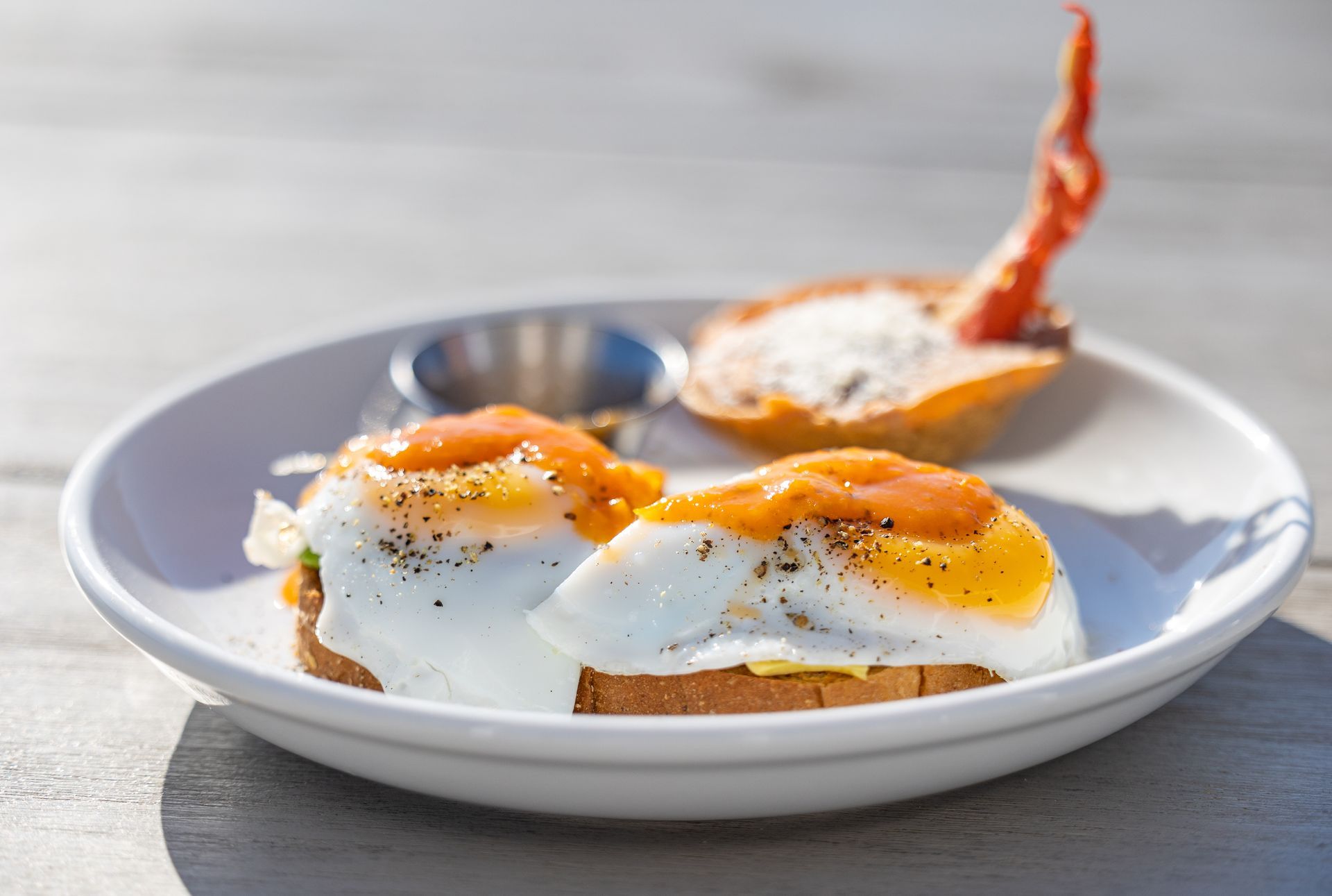 Eggs benedict with orange sauce, side of pastry and small metal cup on a white plate.