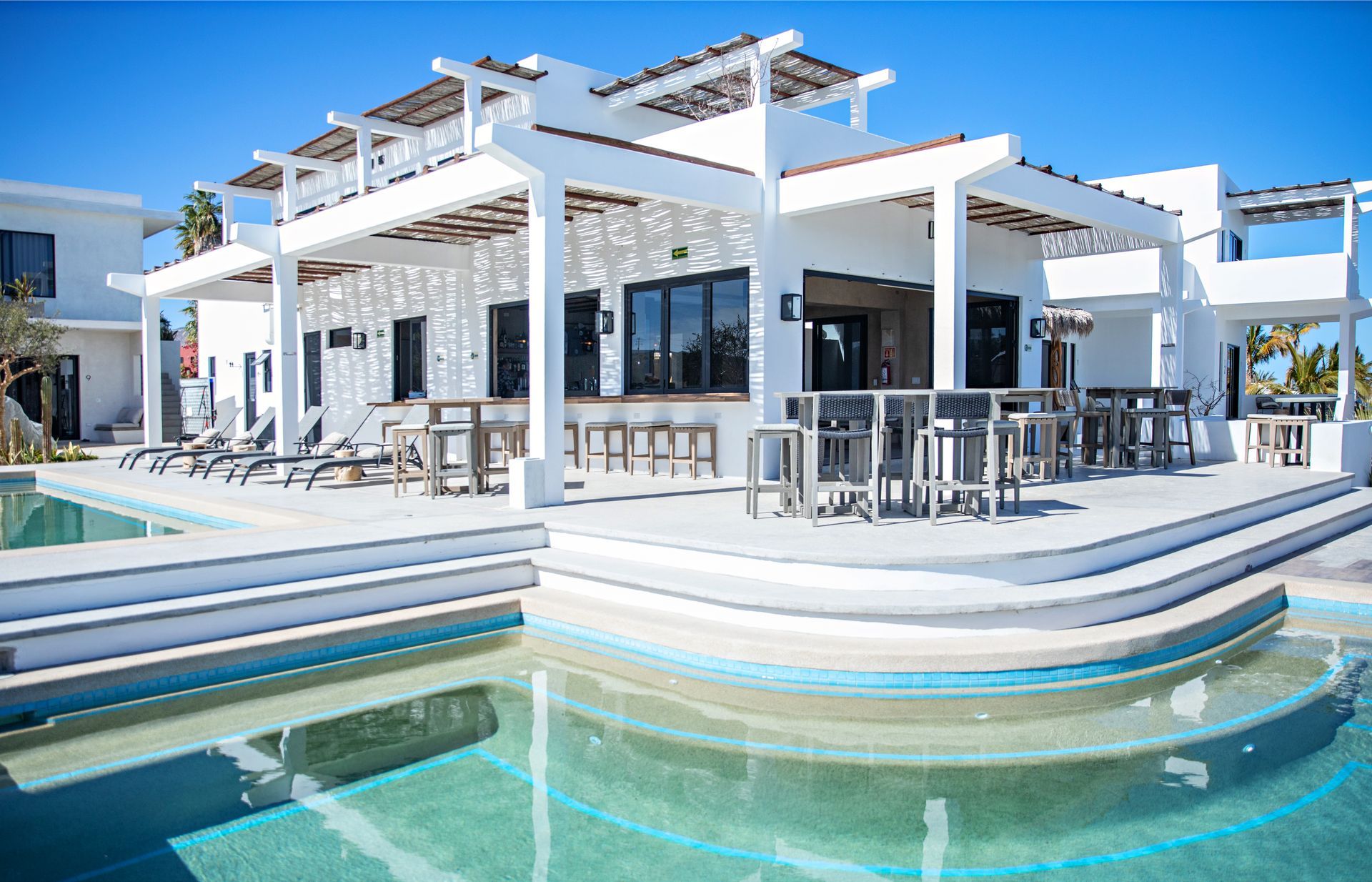 White building with a bar, seating, and pool. Bright blue sky.