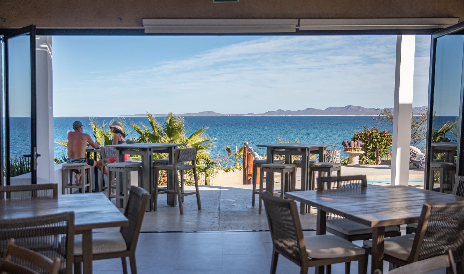 Restaurant patio with ocean view, tables, chairs, people near the bar, blue water, sunny day.