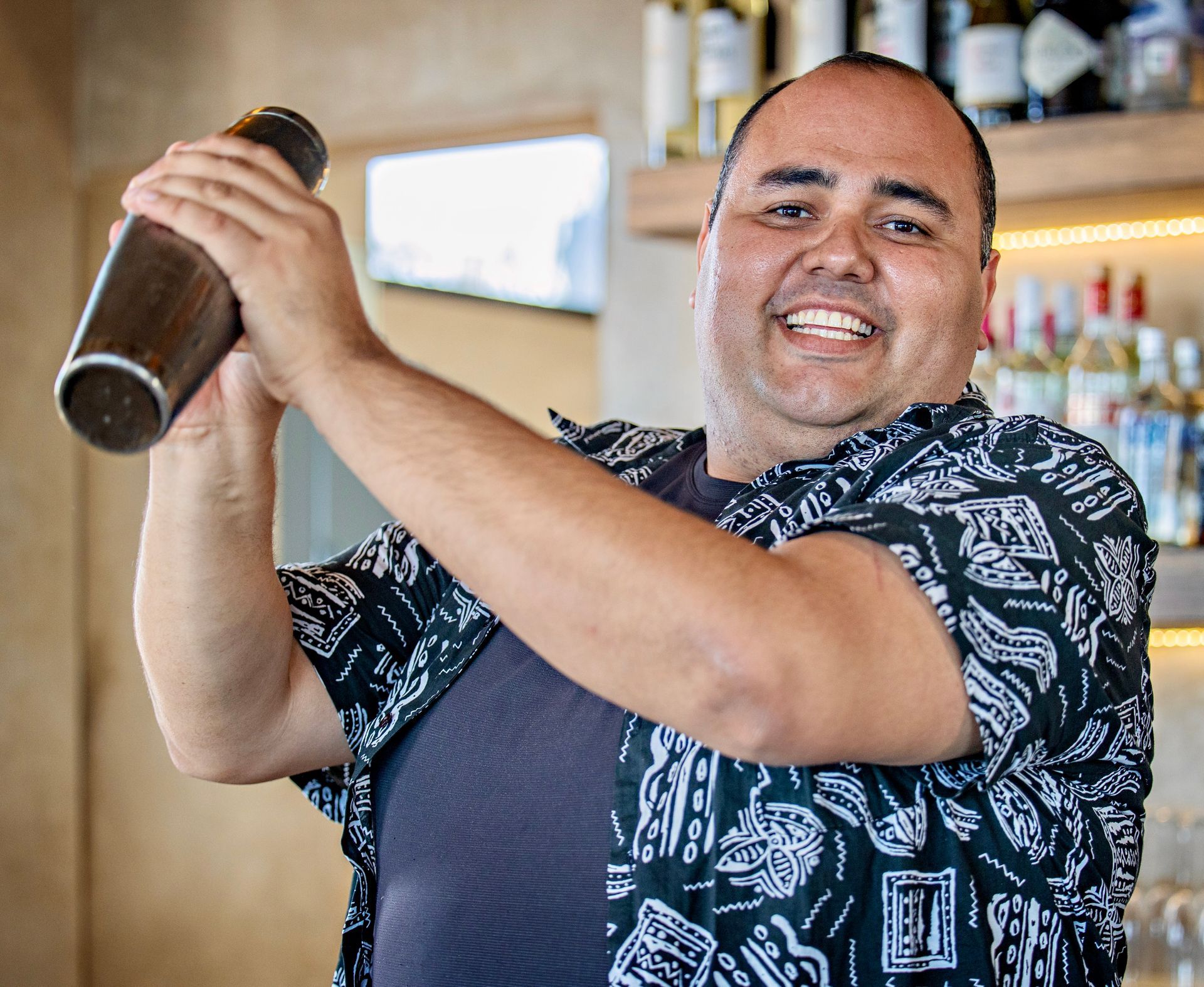 Bartender shaking a cocktail, smiling, in a bar setting, wearing a patterned shirt.