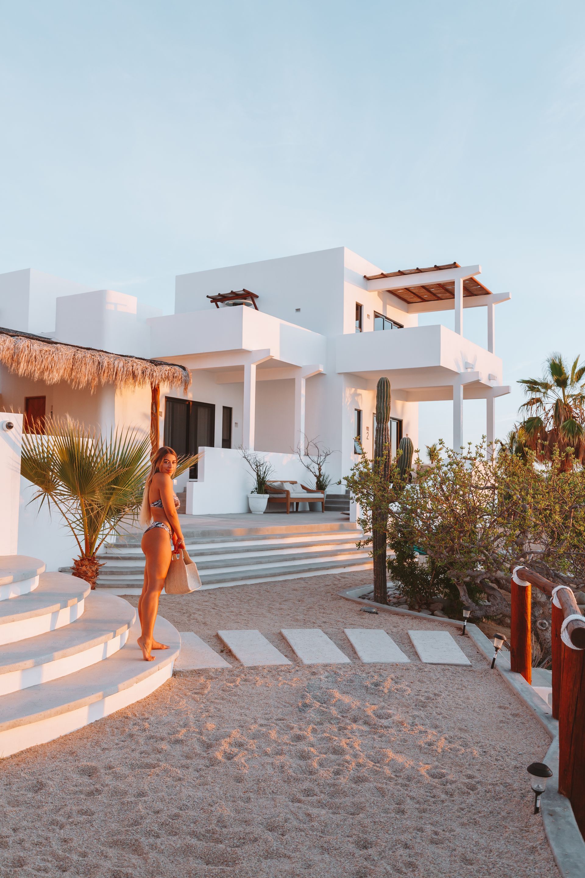 Woman standing near white house with steps, gravel path, and beach bag.